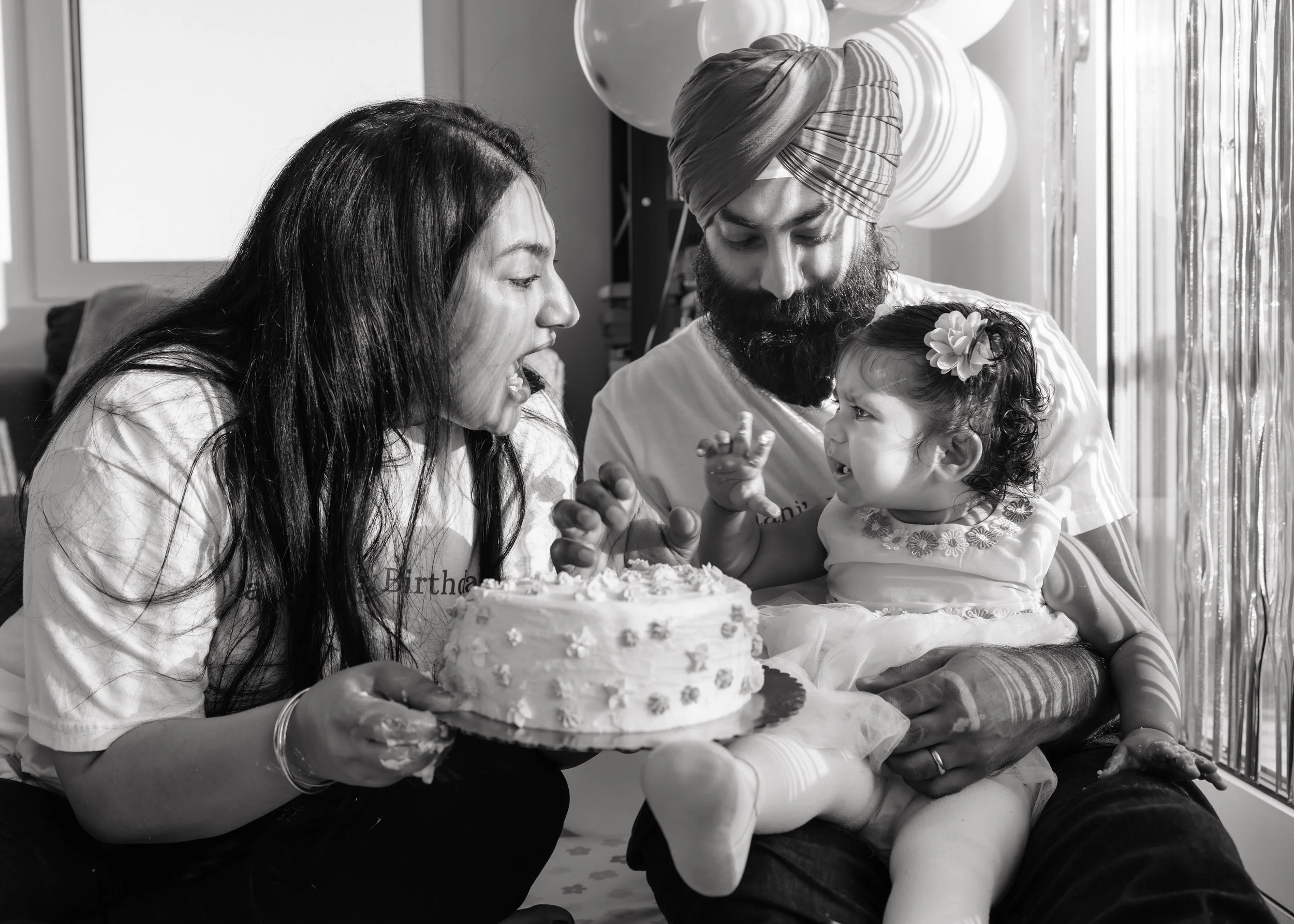 Family celebrating birthday with cake, a woman with long dark hair, a man wearing a turban and a beard, and a young girl with a flower in her hair, indoors with balloons and curtains.