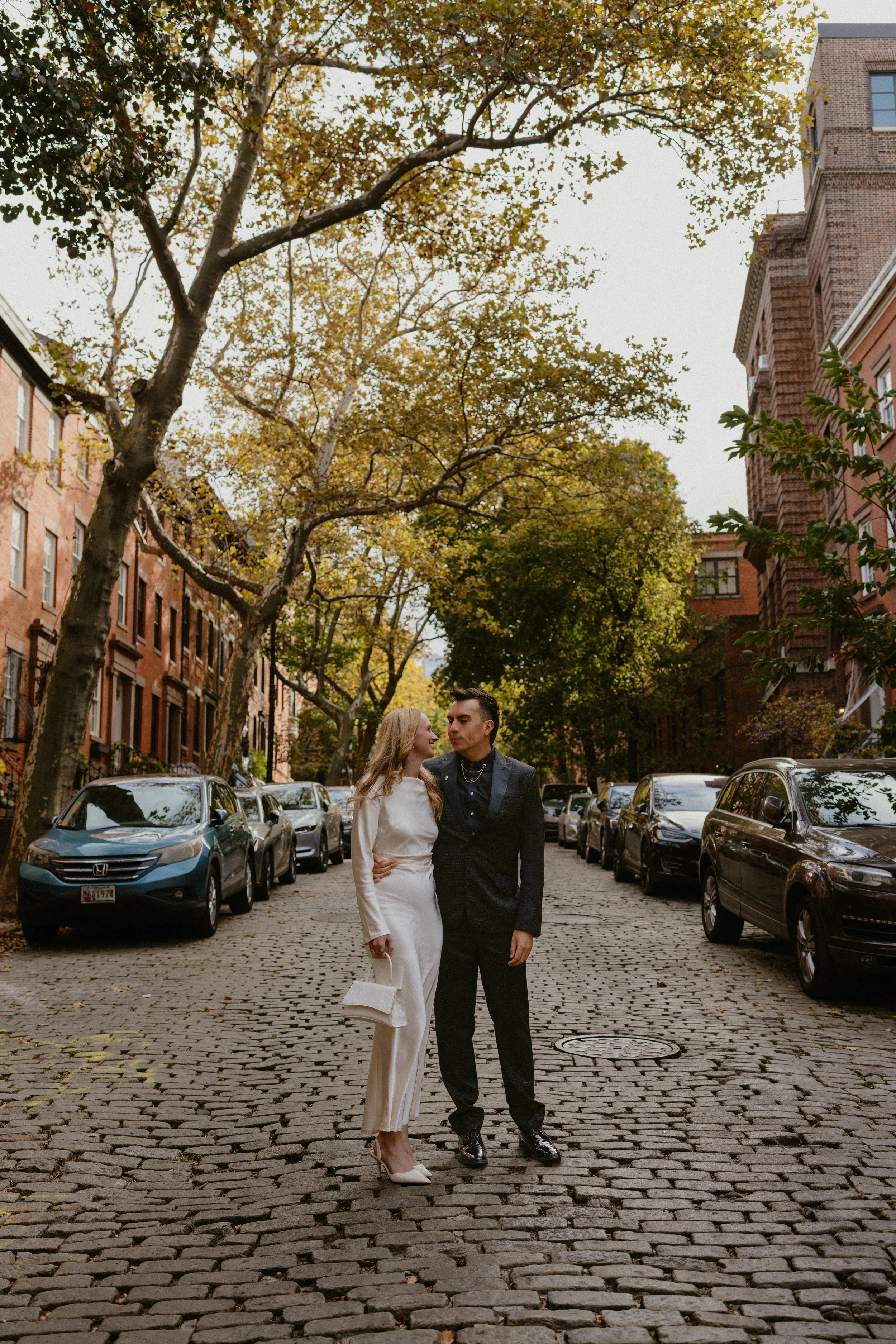 A couple in elegant clothing standing on a cobblestone street lined with parked cars and trees with green and yellow leaves.