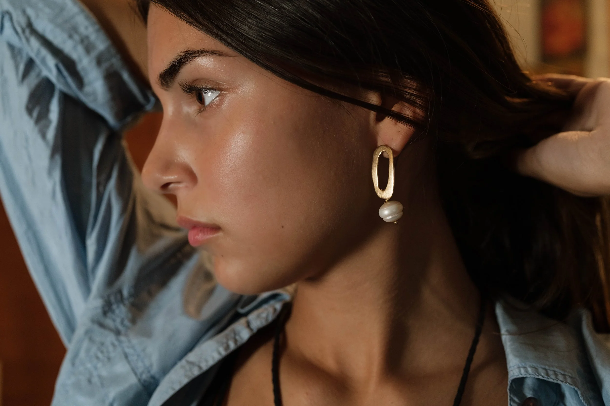 Close-up of a woman with dark hair wearing a large gold and pearl earring, with her arm raised behind her head, wearing a denim shirt.
