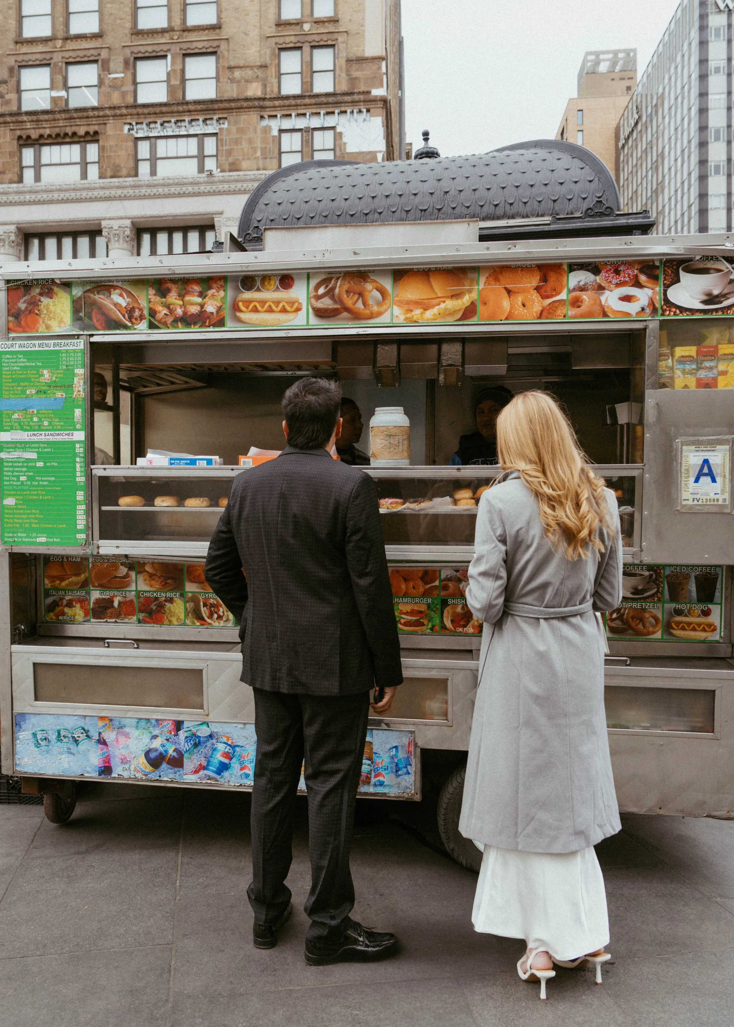 Two people stand in front of a street food truck talking, with the food truck serving breakfast items like sandwiches and donuts, and a poster of various food images on top.