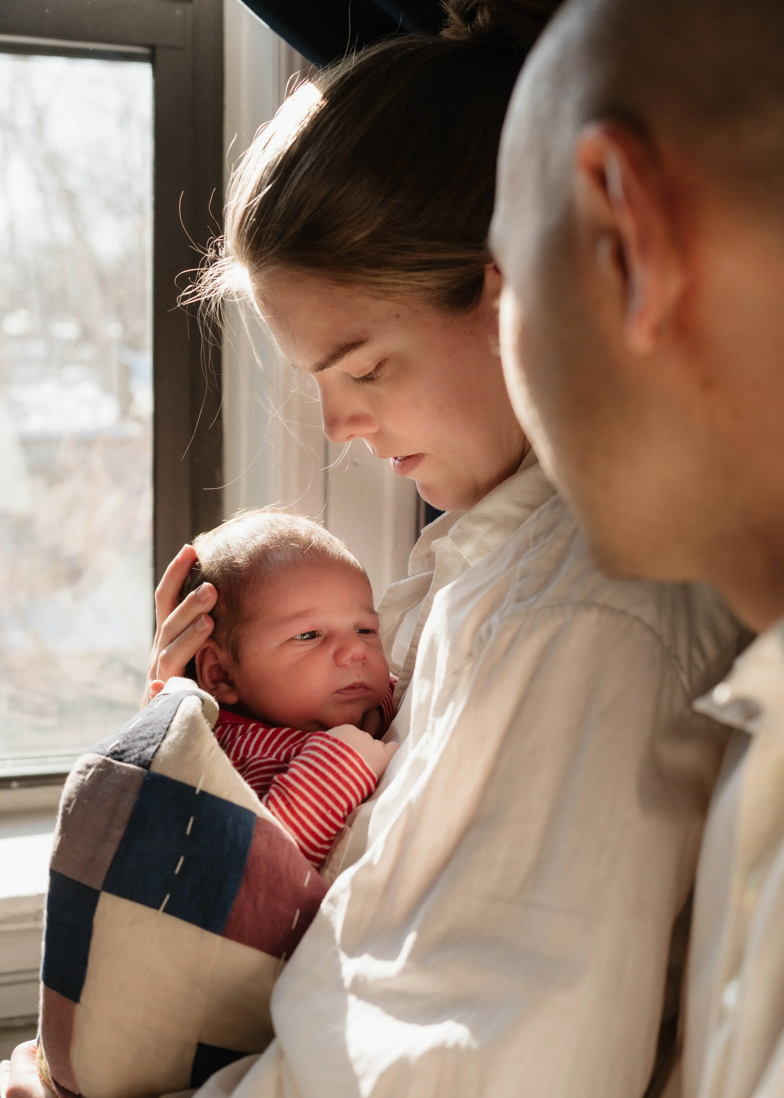 A woman and a man holding a newborn baby by a window, with sunlight streaming in, during daytime.