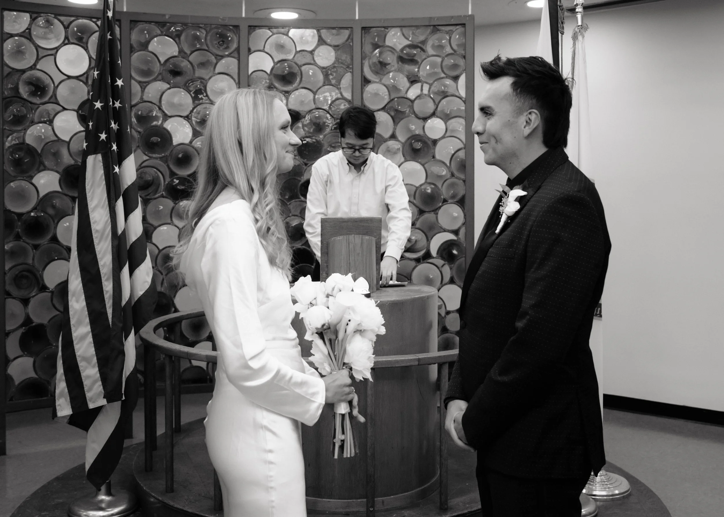 A black and white photo of a wedding ceremony showing a bride and groom facing each other. The bride holds a bouquet of flowers, and a man stands behind a podium with an individual adjusting equipment.
