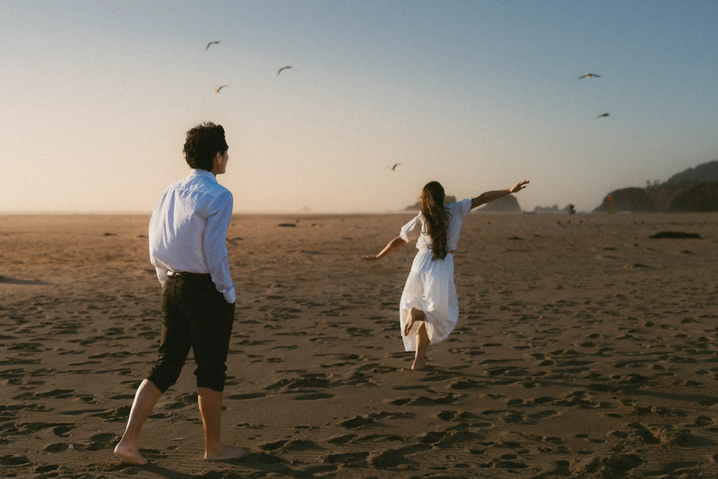 Two people on a sandy beach with one woman running and a man watching, seagulls flying overhead during sunset.