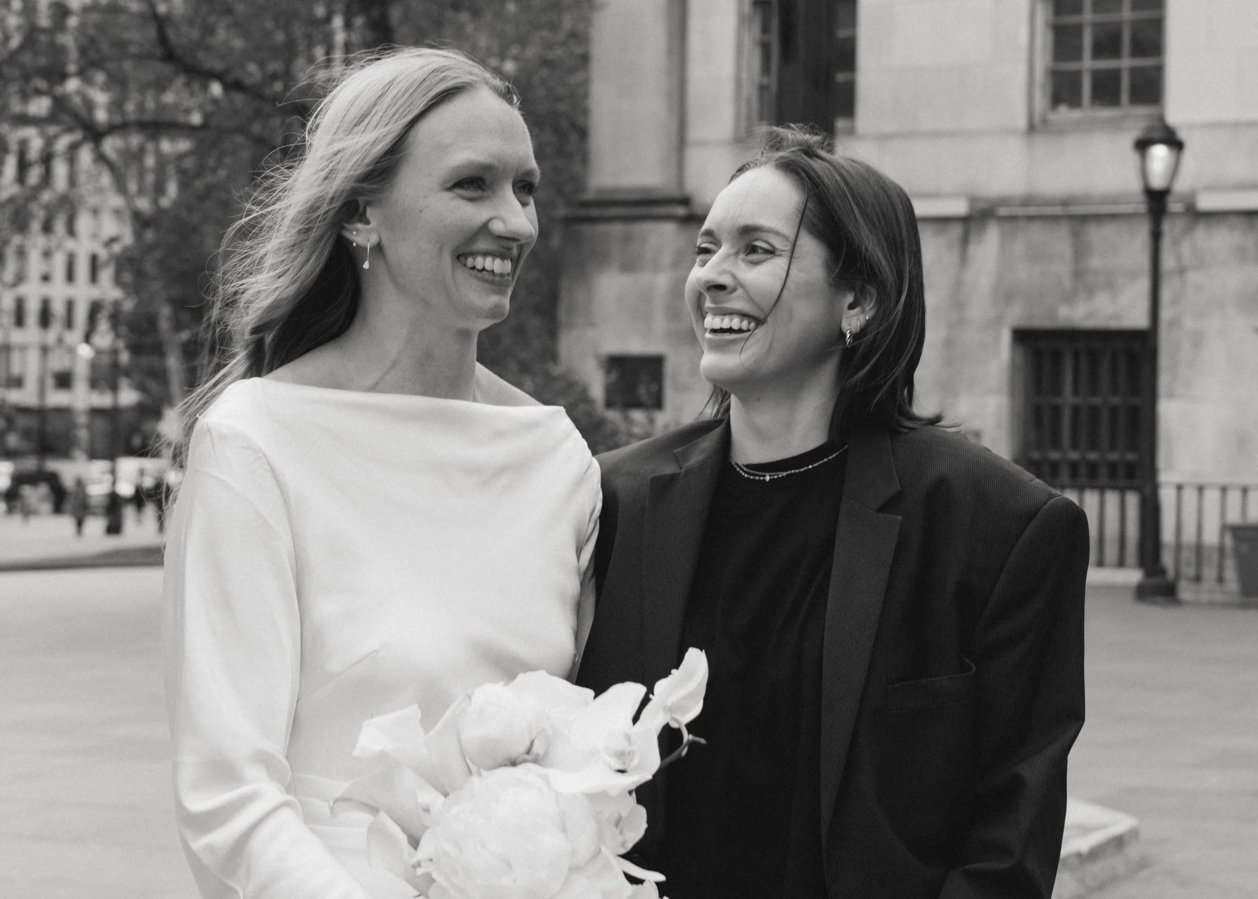 Two women, one holding a bouquet, smiling and looking at each other outdoors.