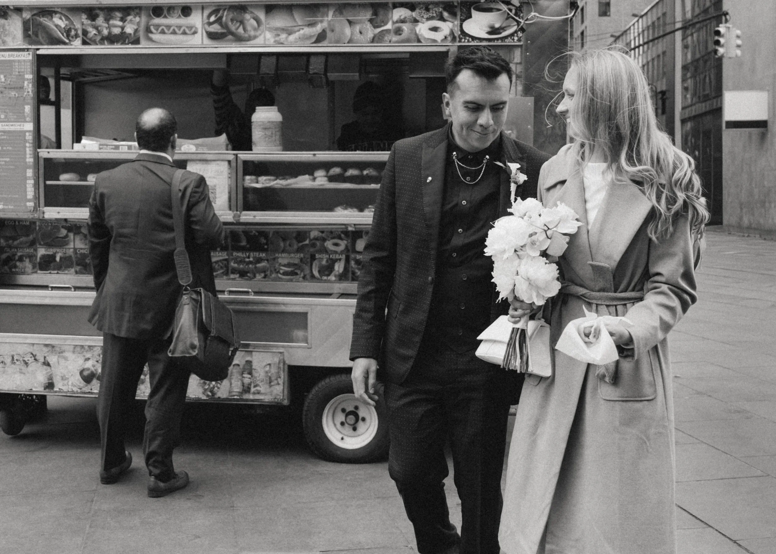 A man and woman walk on city sidewalk near a food cart, the woman holding a bouquet of flowers and the man looking at her, in black and white photo.