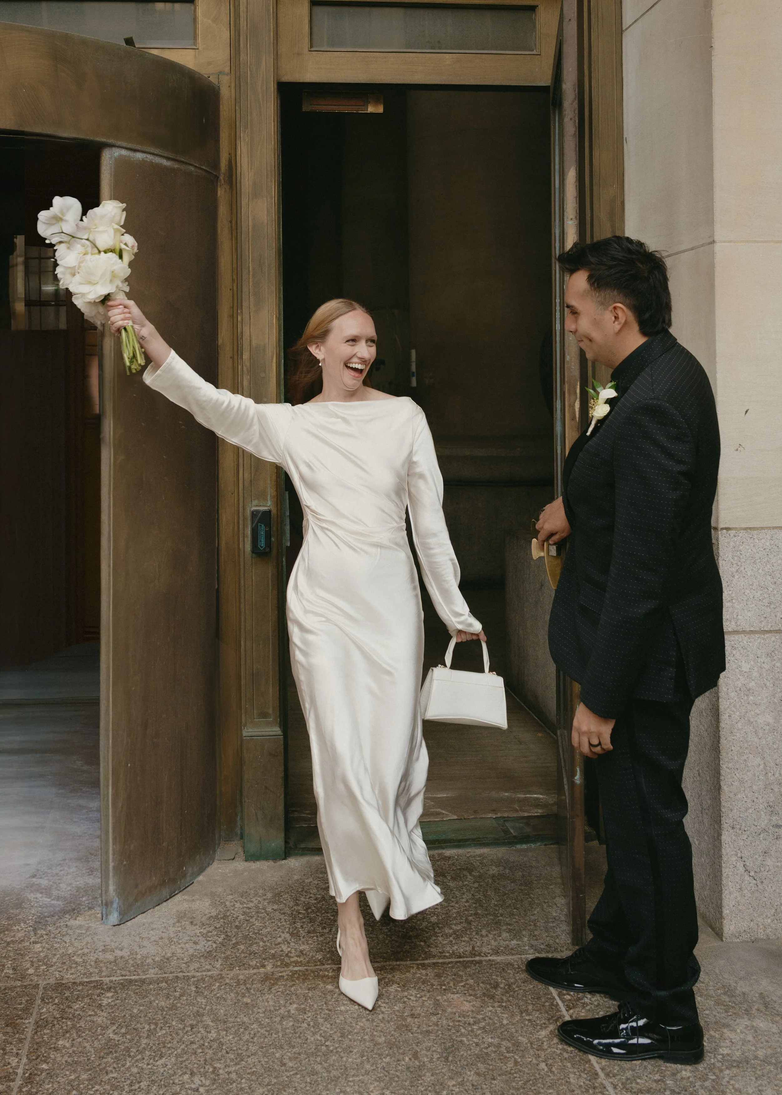 A bride in a white wedding dress and heels joyfully exiting a building holding a bouquet of white flowers, a white handbag, and smiling at a groom in a black suit who is smiling back.