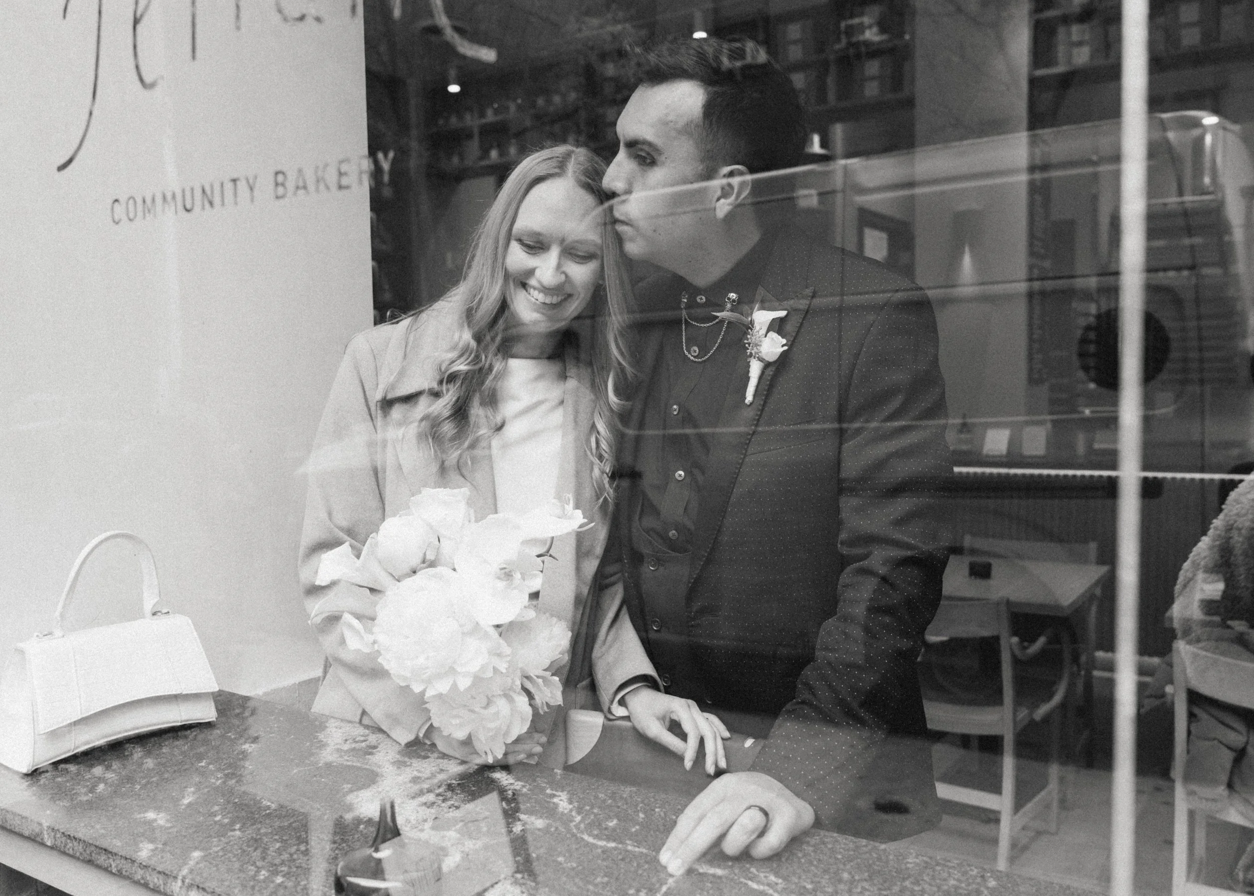 A black and white photo of a couple behind a bakery display window, with the woman holding a bouquet of flowers and smiling, and the man kissing her on the head.
