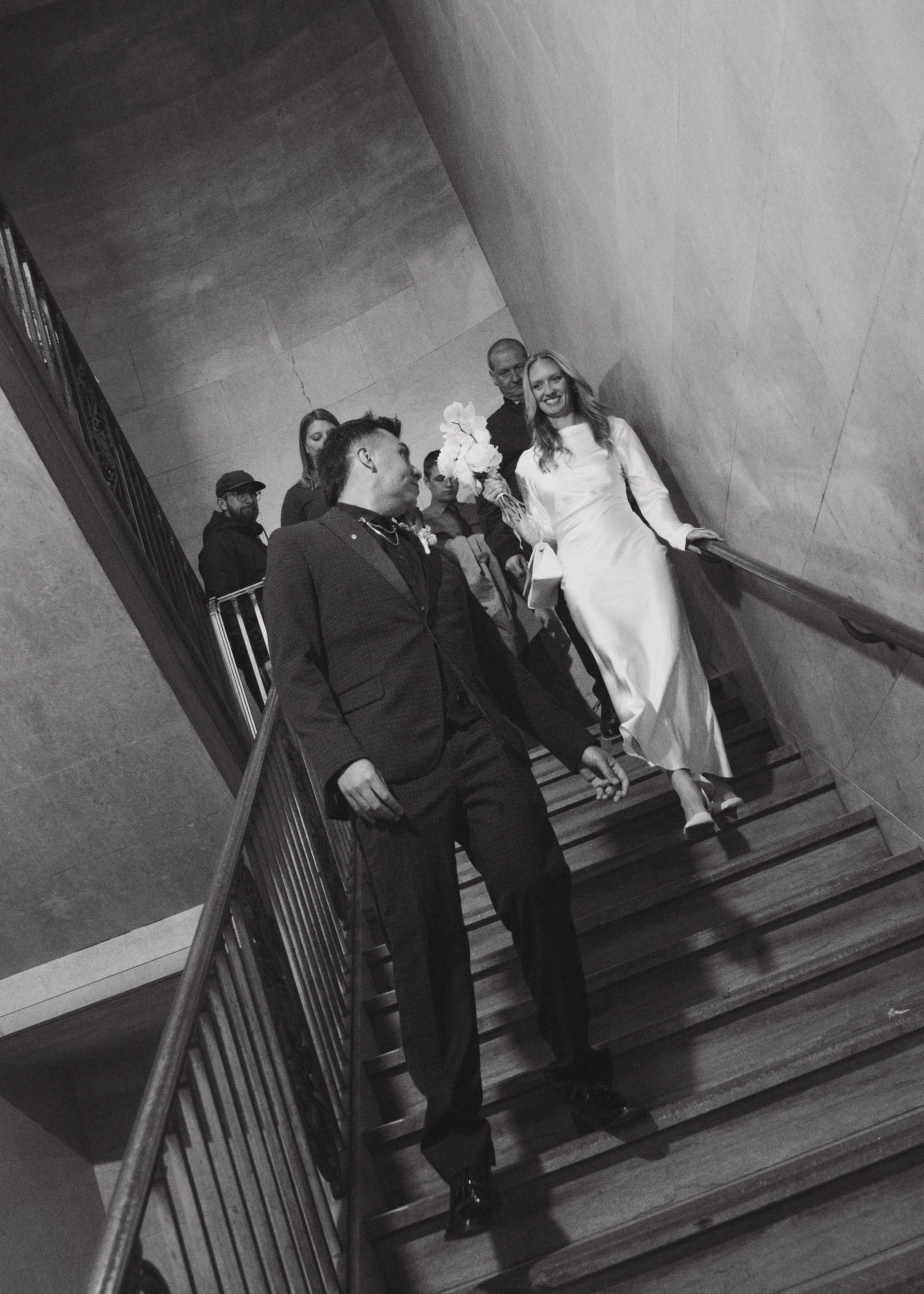 A black-and-white photo of a bride in a long white dress holding a bouquet, walking down a staircase with a group of people, including a man in a tuxedo leading her, in an indoor setting with plain walls.