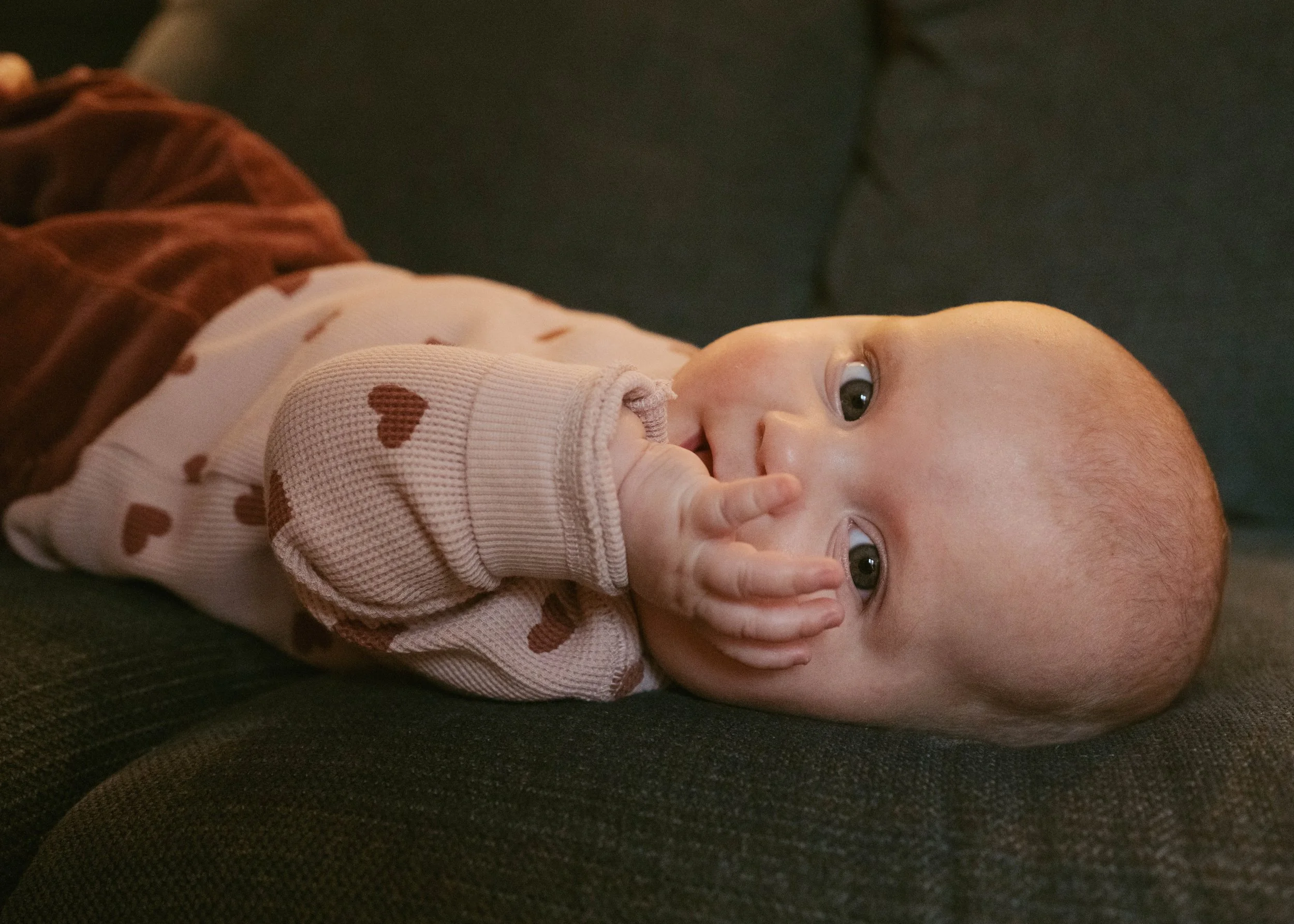 A baby lying on a dark couch, wearing a pink sweater with brown hearts and brown pants, with one hand in their mouth and big, bright eyes looking at the camera.