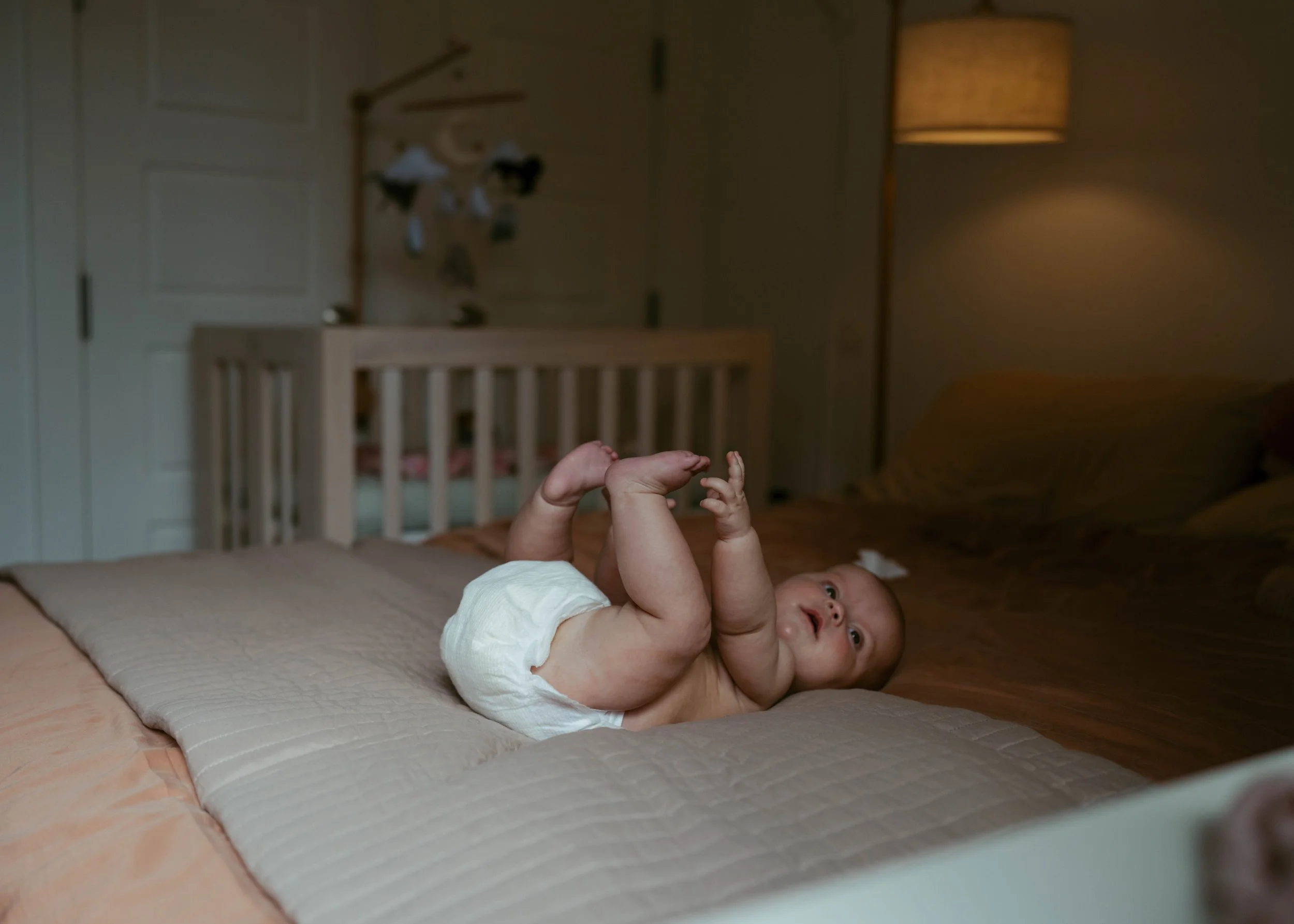 A baby lying on a bed in a dimly lit room, wearing only a diaper, with legs bent and hands reaching towards the feet, looking up.