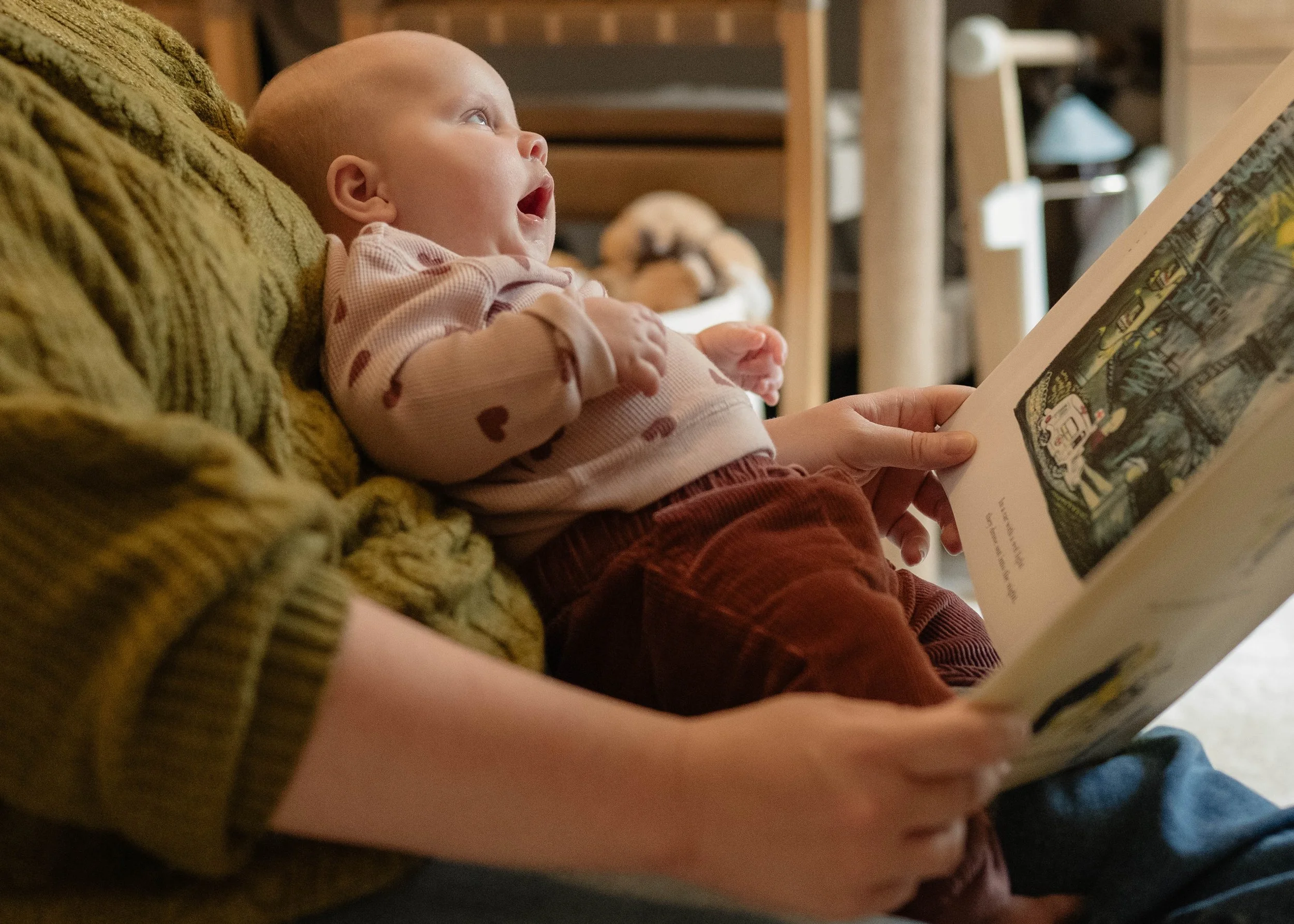 A young child with a bald head lying on a green knitted blanket, looking up and reacting with curiosity while being read a book by an adult who is holding the book. The scene is indoors, with a cozy background including wooden furniture and a lamp.