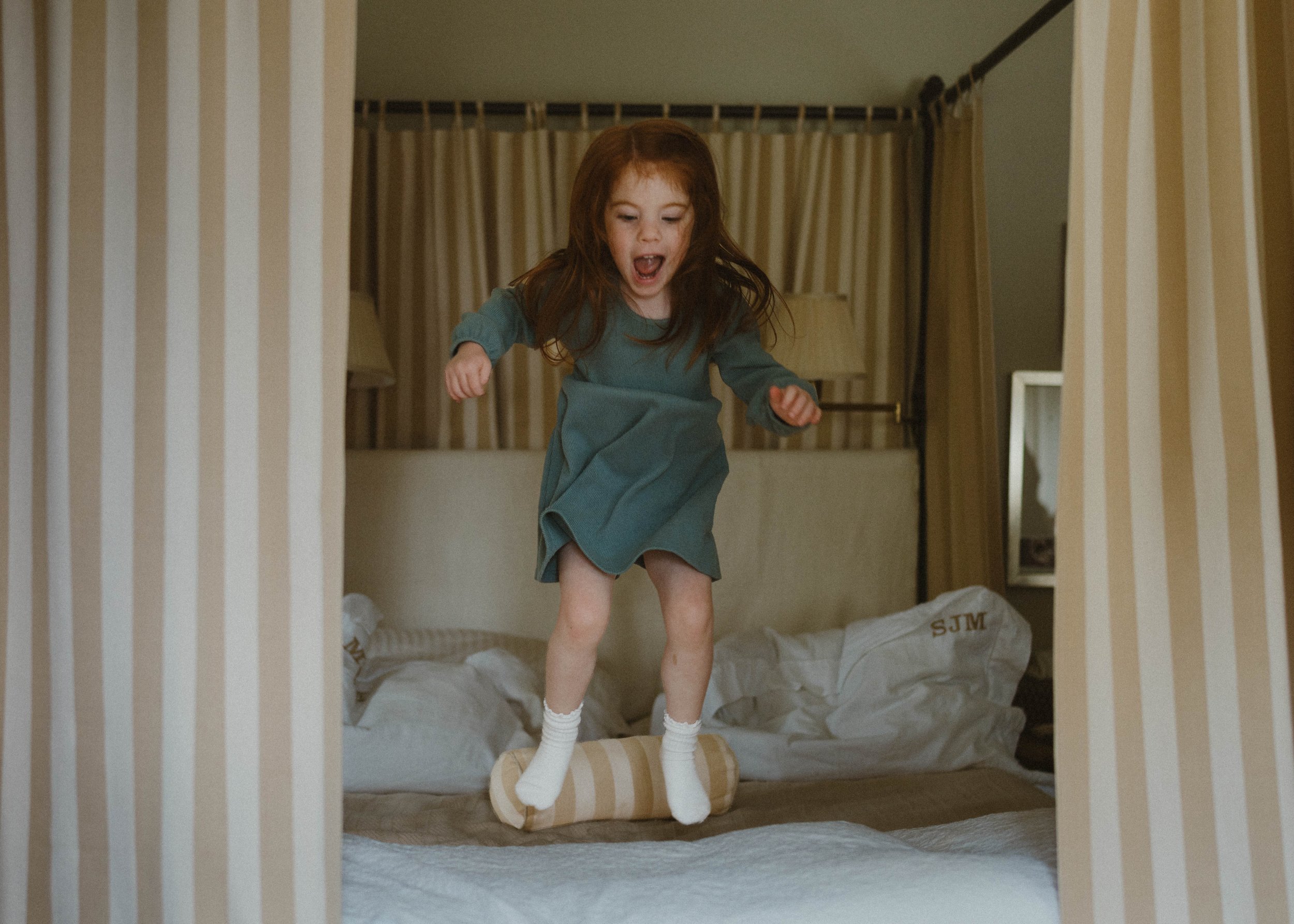 A young girl with long, reddish hair, wearing a blue long-sleeve dress, white socks, and no shoes, standing on a small wooden stool on a bed with bedding, in a bedroom with beige curtains and bags on the floor.