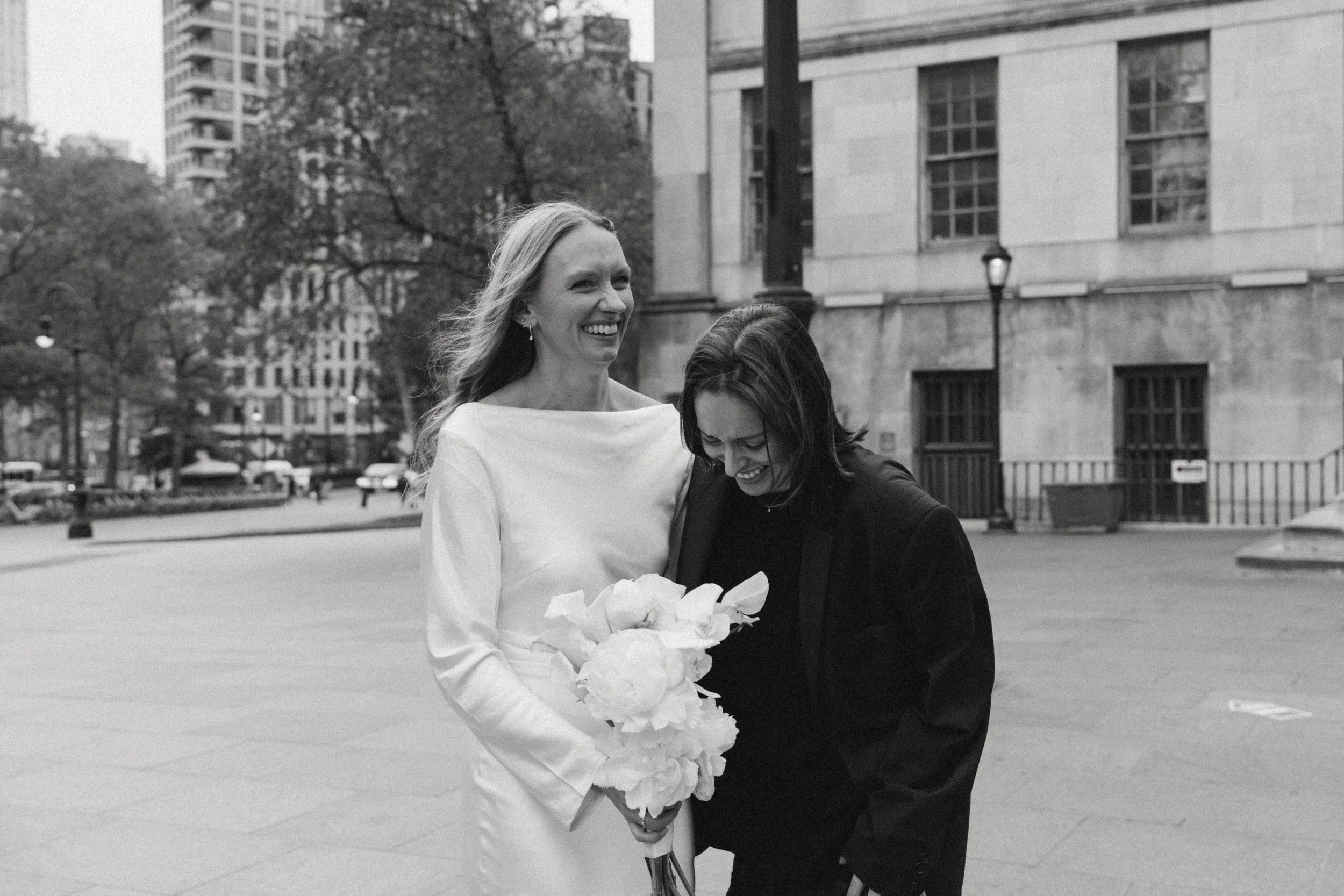 Two women, one in a white dress and the other in black, smiling and sharing a laugh, with one holding a bouquet of flowers, on an urban street in black and white.
