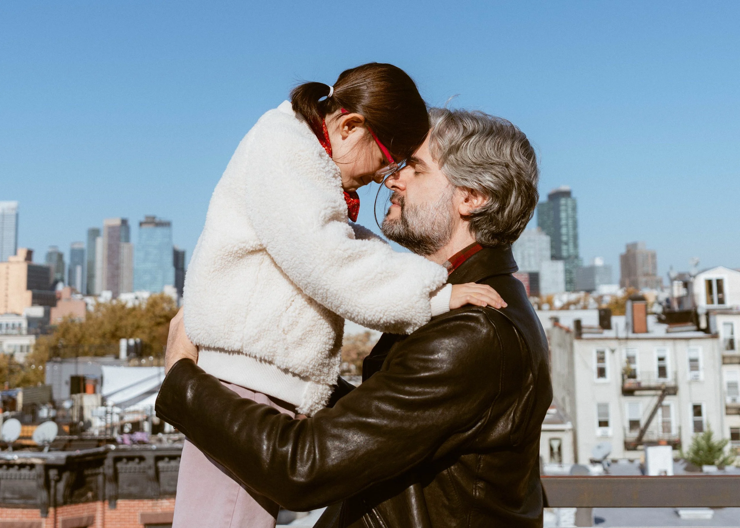 A man lifts a woman in a white fuzzy jacket and glasses, touching foreheads with her against a city skyline background on a clear day.