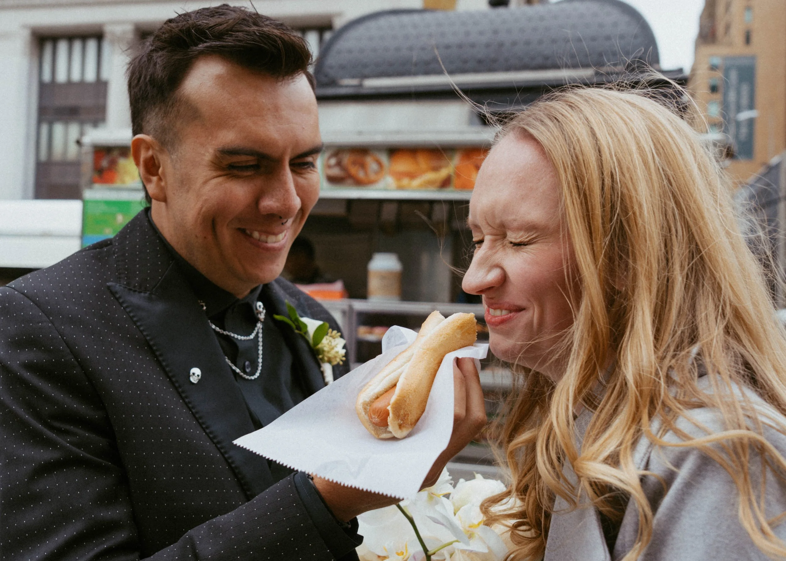 A man and woman share a funny moment as the man holds a hot dog up to the woman's face, which she is about to bite. The man is smiling with his eyes closed, and the woman has her eyes scrunched with her nose wrinkled in anticipation. They are outdoor