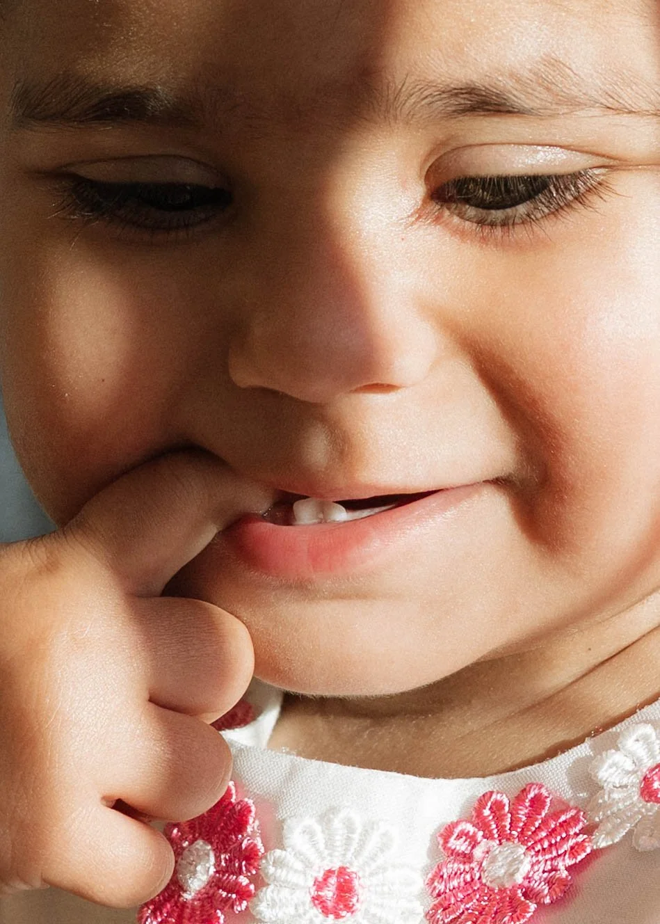 A close-up of a young child's face with one finger on their lip, showcasing a loose front tooth and wearing a white garment with pink and white floral embroidery.