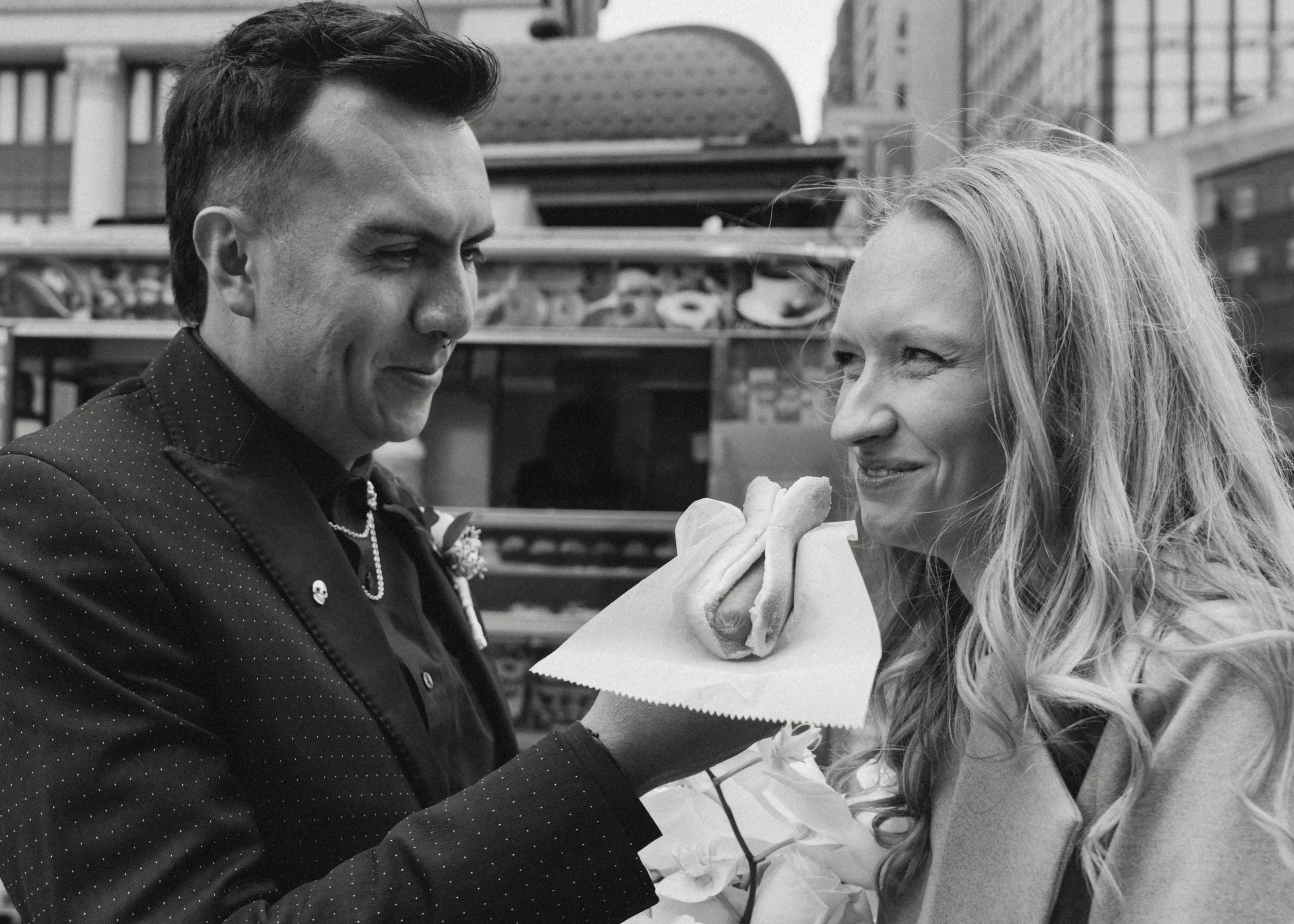 A man holding a hot dog in front of a woman outdoors. The man is wearing a dark, polka-dotted jacket with pins and a necklace, and the woman has long, wavy hair and is smiling as she looks at the hot dog.