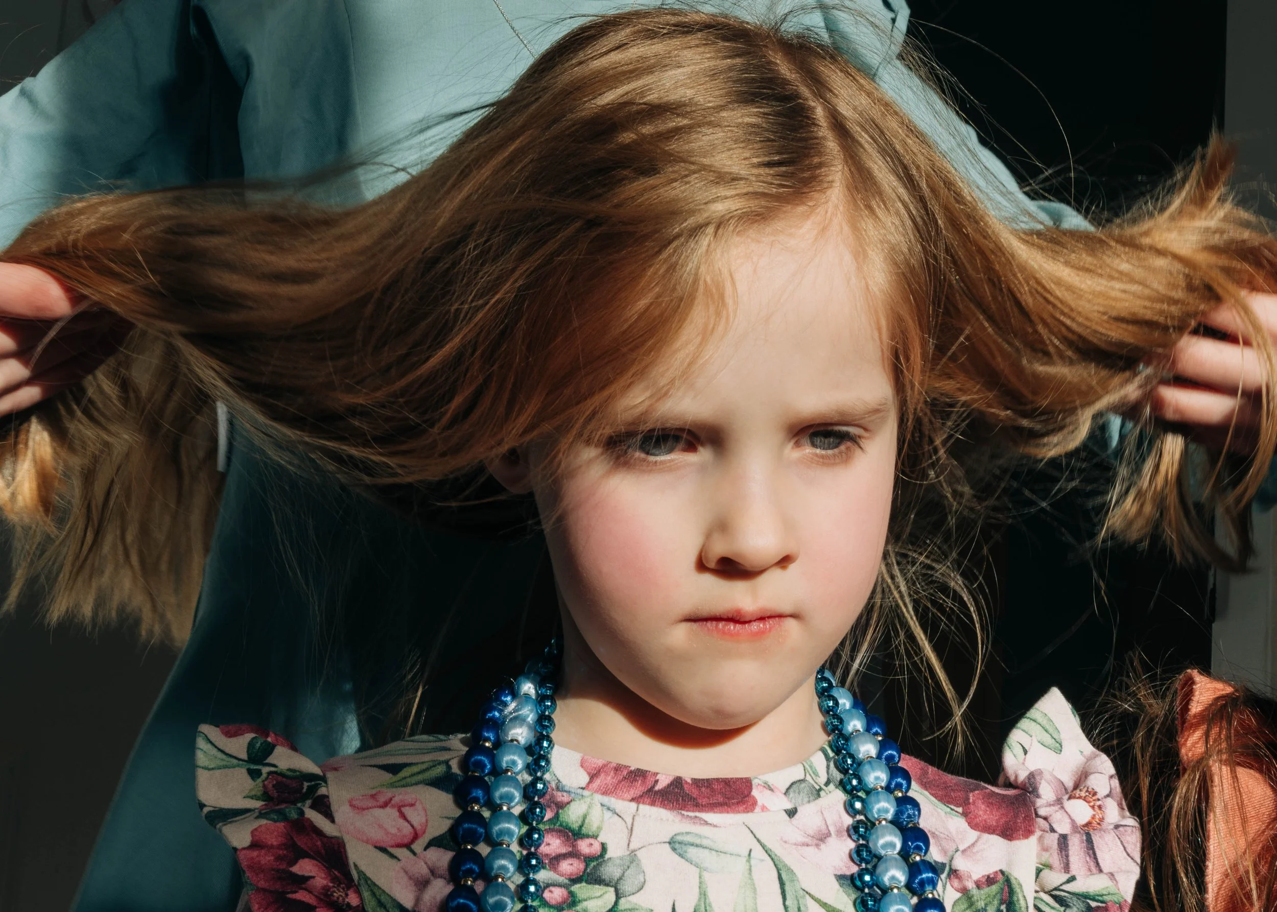 A young girl with long red hair, wearing a floral dress and blue pearl necklaces, holds her hair out to the sides and looks downward with a serious expression.