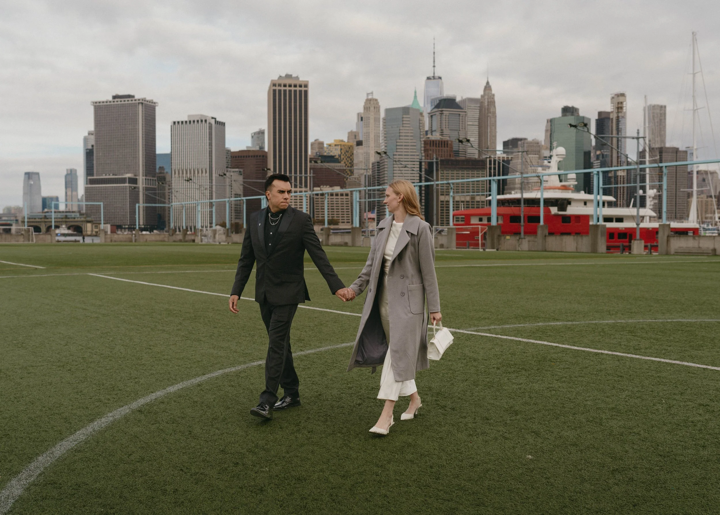 A man and woman holding hands and walking on a sports field in Brooklyn, New York, with the Manhattan skyline in the background.