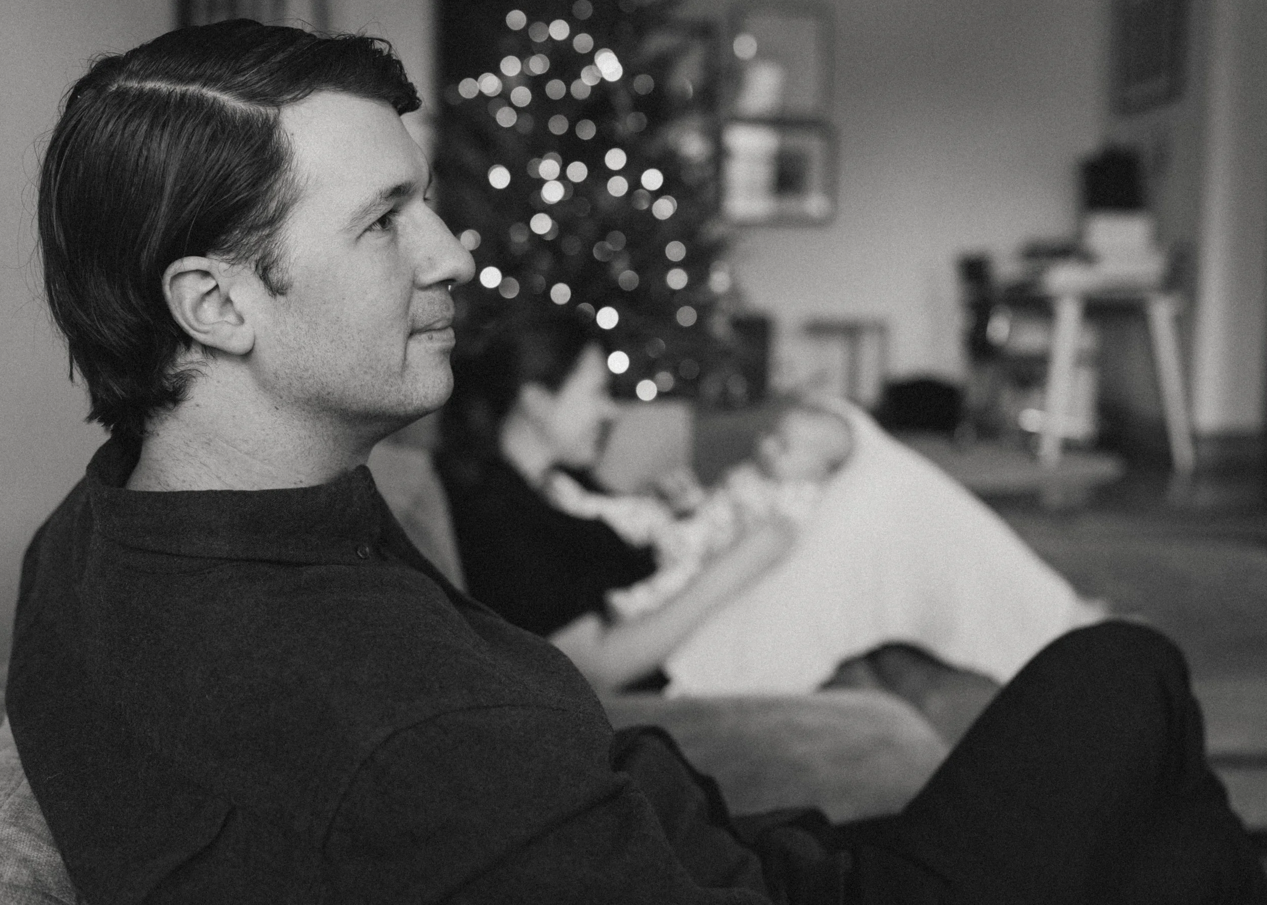 A man with dark hair sitting on a couch, gazing to the left, with a Christmas tree and a woman with a baby in the background, in a living room decorated for Christmas.