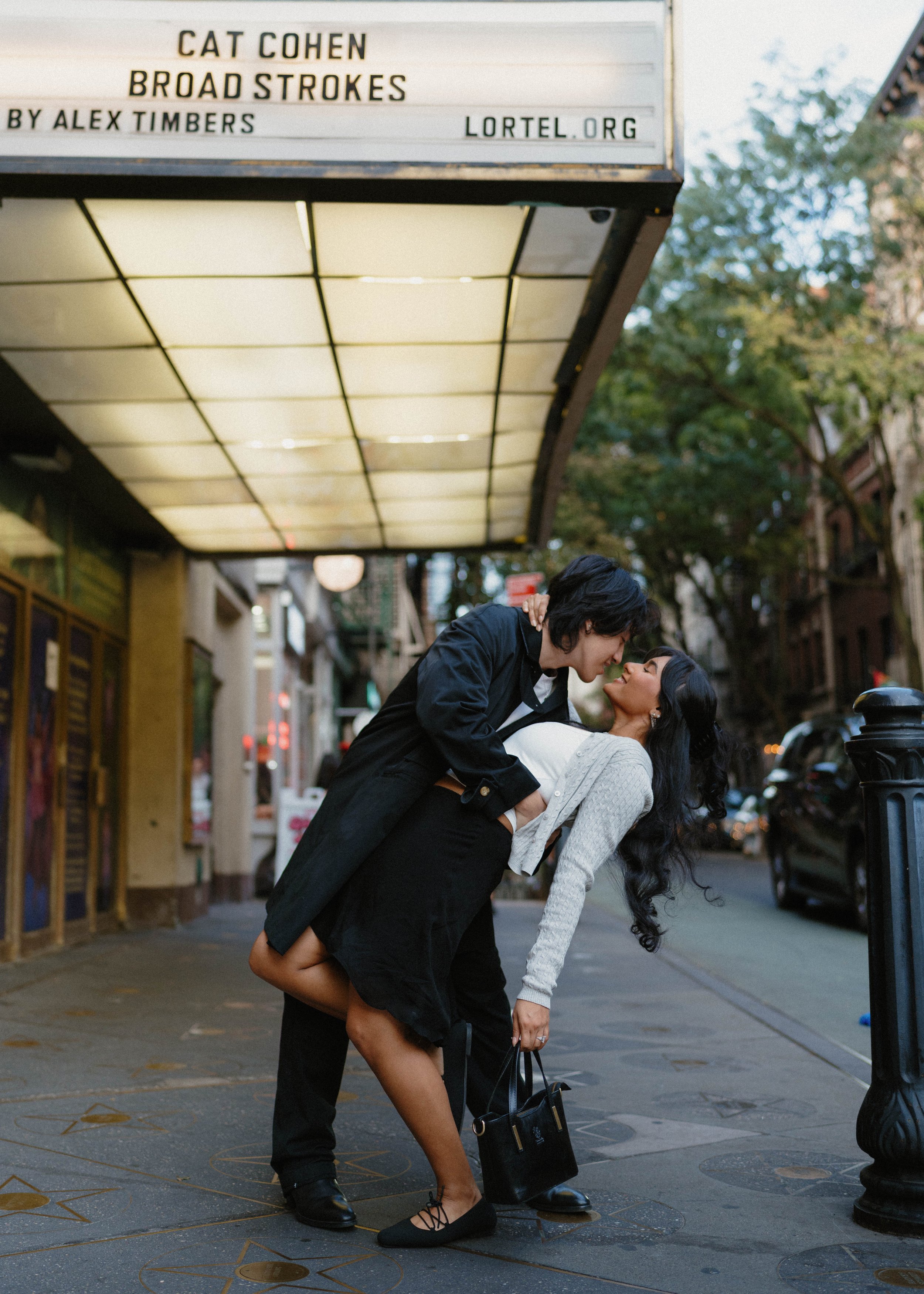 A man and a woman dance closely on a city sidewalk, with the man leaning over the woman who is bending backward. They are outdoors near a marquee with a sign that reads 'CAT COHEN BROAD STROKES BY ALEX TIMBERS LORTEL.ORG.' The woman is holding a black handbag, wears black shoes, and has long dark hair; the man wears dark clothing and black shoes.