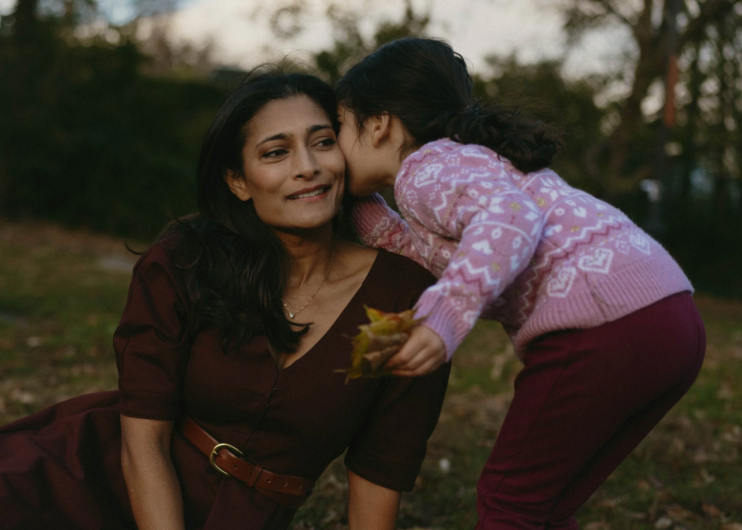 A woman in a maroon dress lying on the ground with a slight smile, while a young girl leans over, whispering into her ear and holding a few yellow leaves, in an outdoor park setting during autumn.