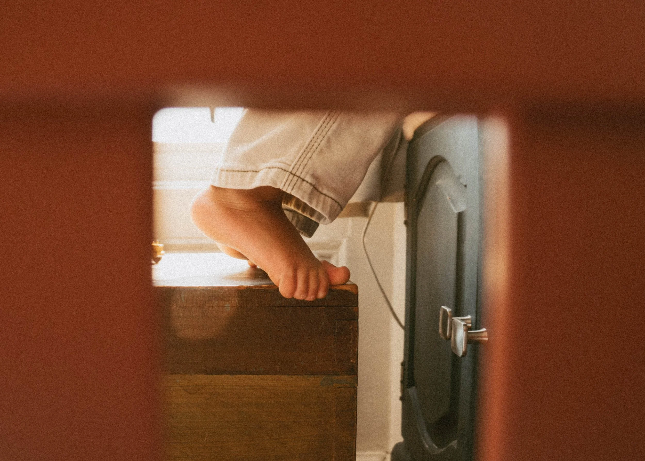 Child standing on a wooden surface, with only legs visible, wearing khaki shorts and black shoes, viewed through a rectangular opening.