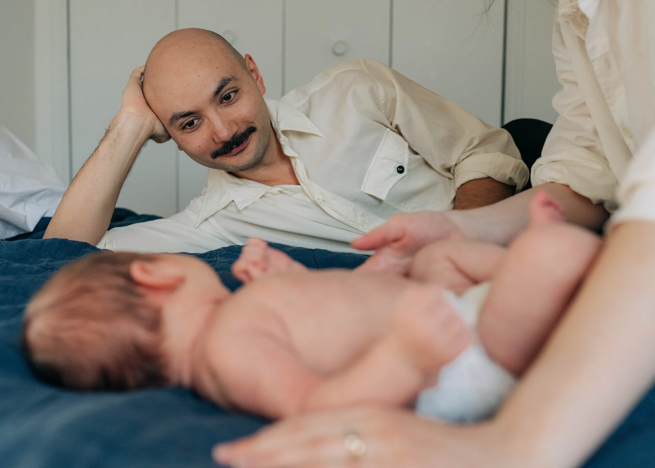 A man with a bald head and mustache lying on a bed, looking at a newborn baby lying on the bed beside him.
