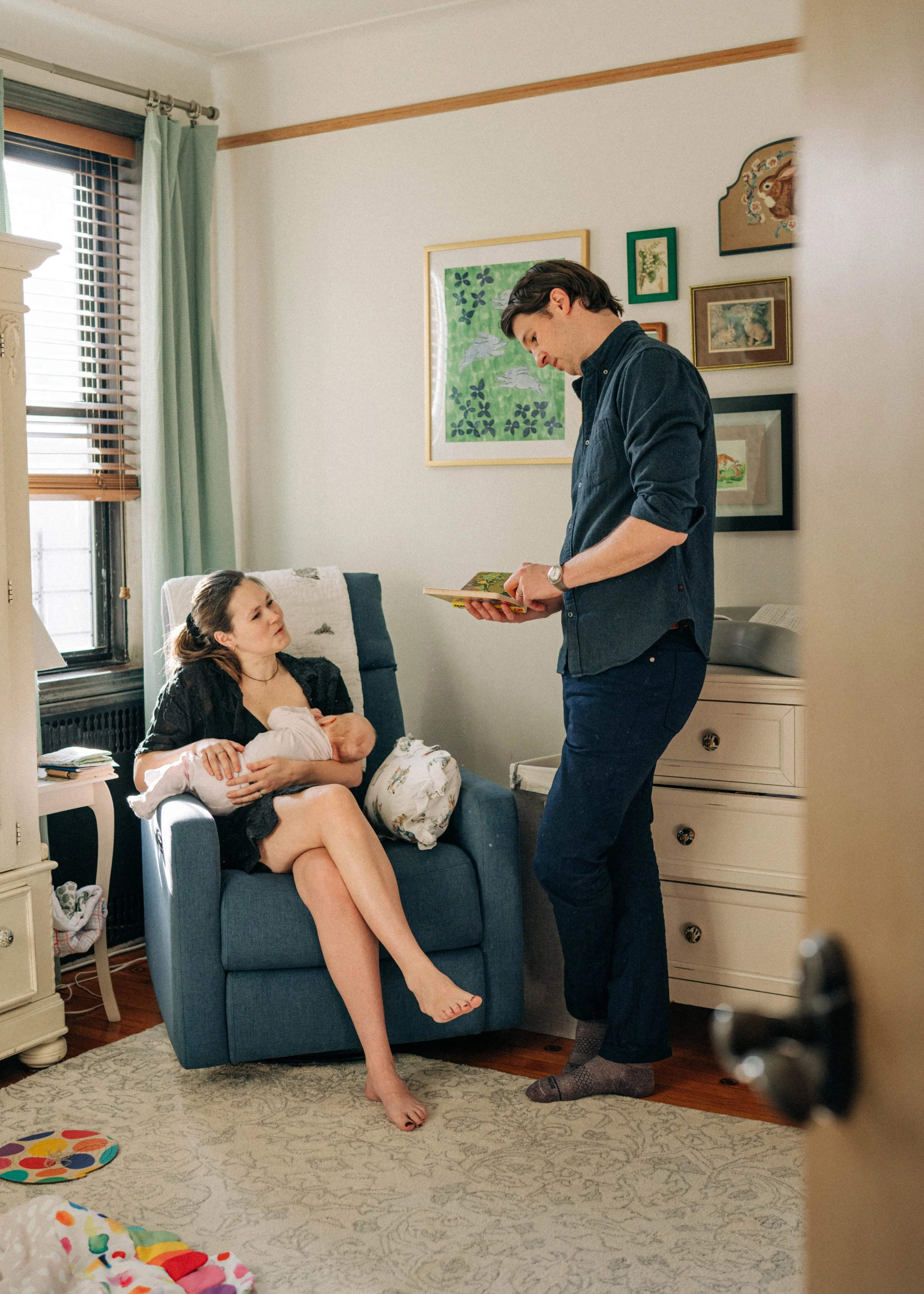 A woman sitting in a blue armchair holding a newborn baby, looking up at a man standing in front of her reading a book, inside a cozy living room with framed artwork on the wall and a window with blinds.