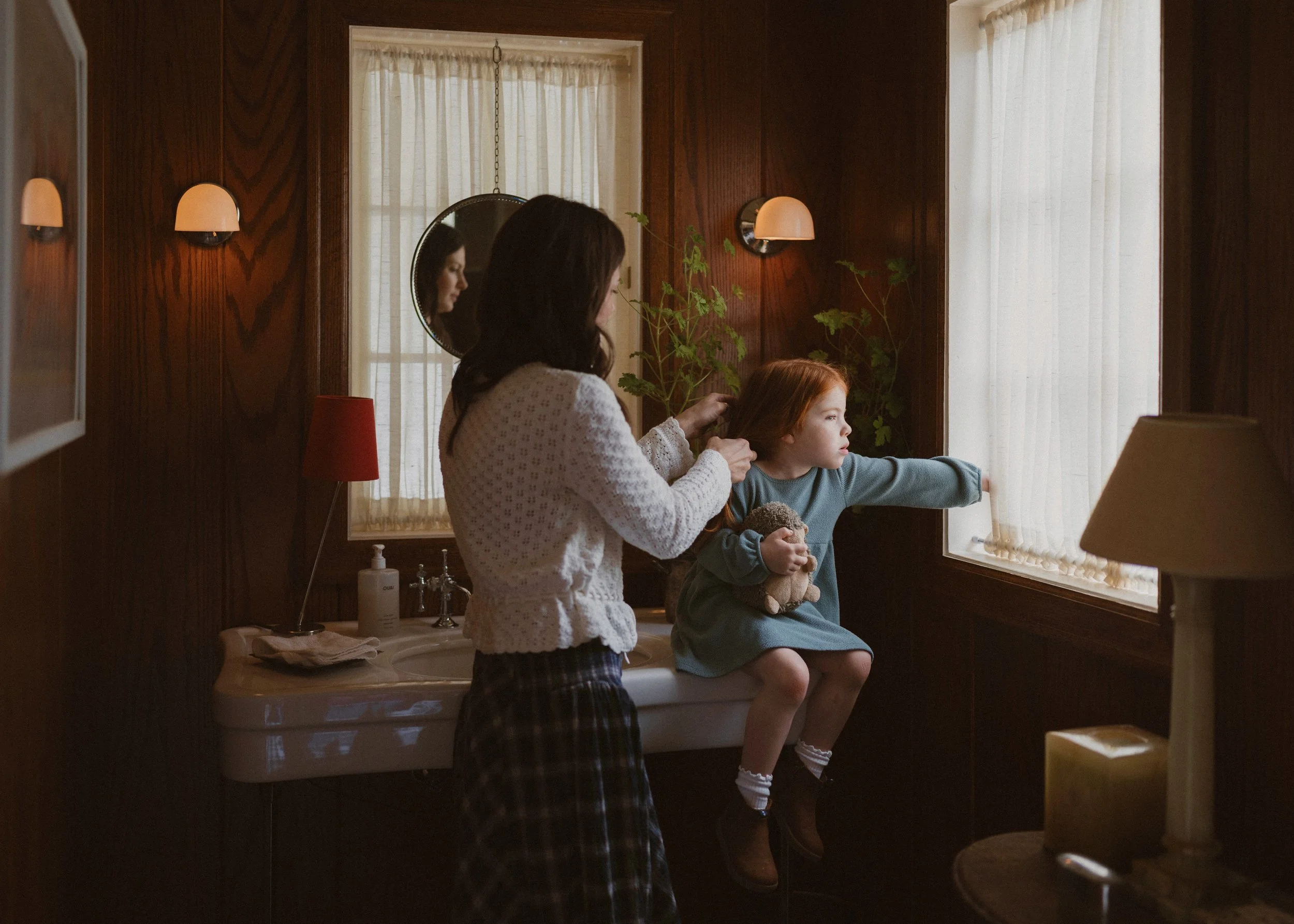 A young woman with dark hair and a young girl with red hair are in a bathroom. The girl is sitting on a sink, pointing out a window, while the woman does the girl's hair. The bathroom has wood-paneled walls, two wall sconces, a mirror, and a plant ne
