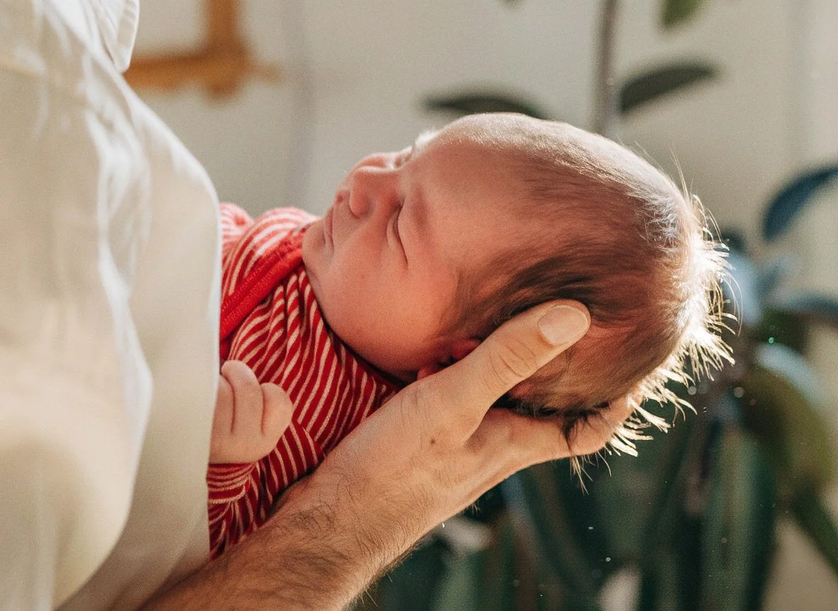 An adult holding a sleeping baby's head with one hand, cradling the baby's head close to their chest, in a cozy setting.
