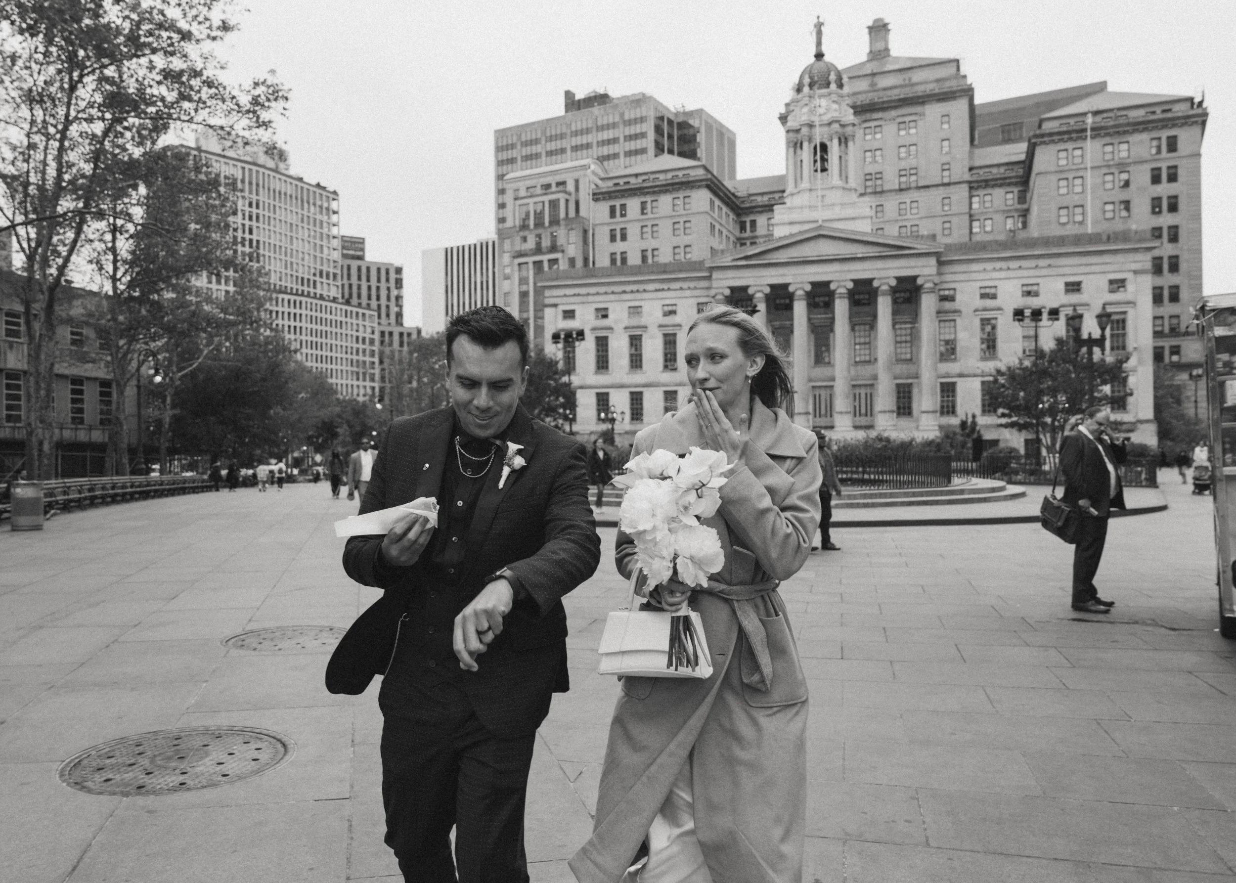 A black and white photo of a man and woman dressed formally, walking in an urban area. The man is holding a small item and looking at his watch, while the woman, holding a bouquet of flowers and a purse, is looking at him with her hand near her face.