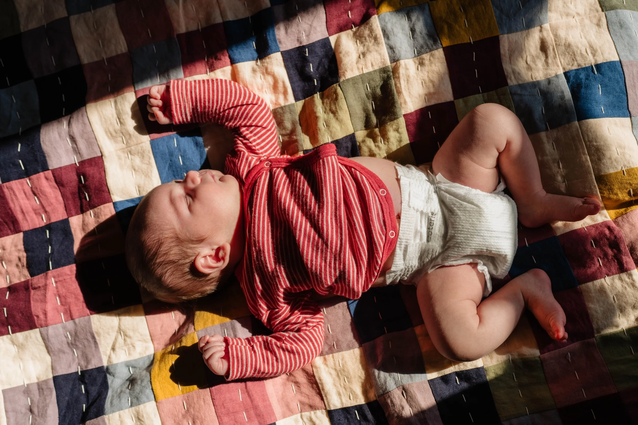 A baby sleeping on a multicolored patchwork quilt, wearing a red and white striped long-sleeve shirt and a diaper.