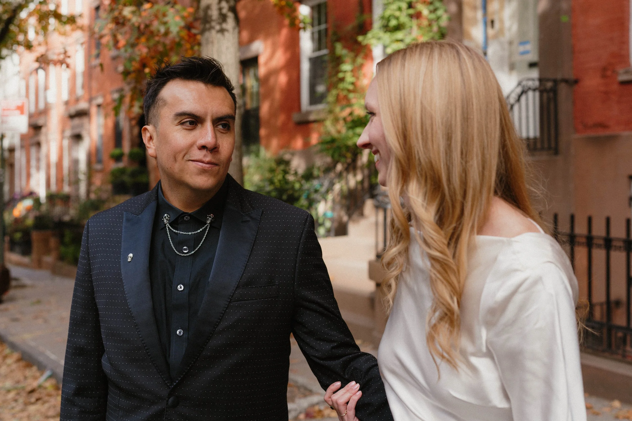A man and woman stand outdoors on a sidewalk in a residential neighborhood, looking at each other and smiling. The man is dressed in a black suit with a chain necklace, and the woman is wearing a cream-colored blouse with long, light red hair.