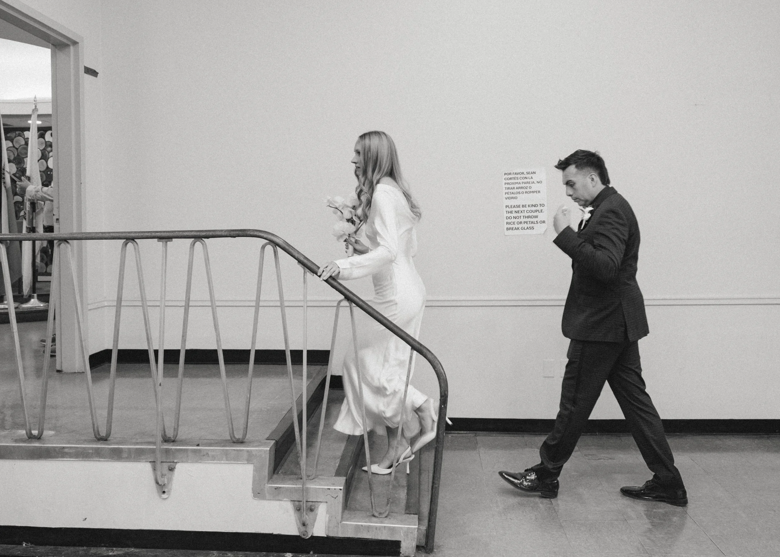 A black and white photo of a woman in a wedding dress ascending a staircase and a man in a suit following her, both indoors. The woman has long hair and is carrying a bouquet of flowers, while the man is looking down at his hand. There is a sign on t