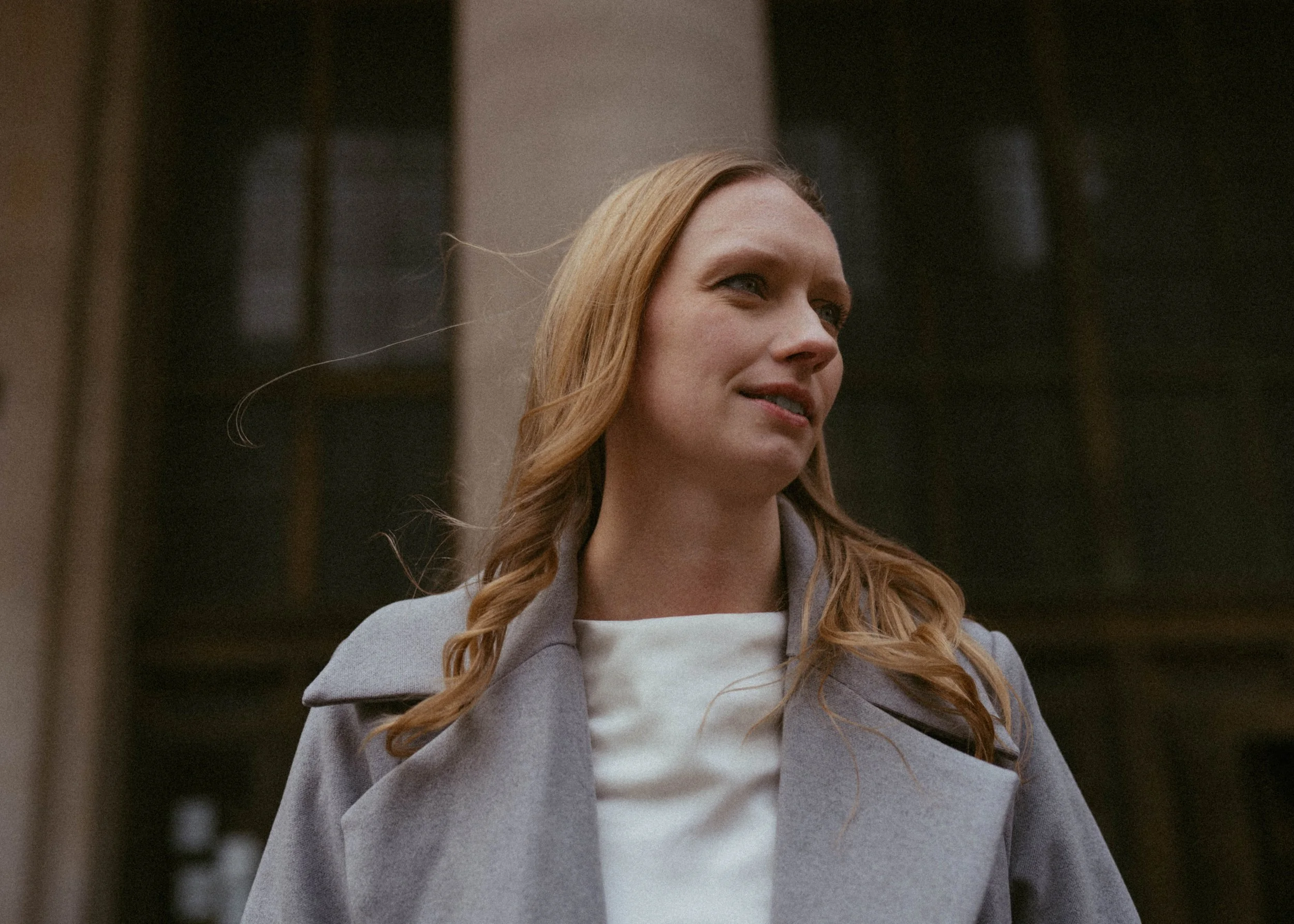 A woman with long red hair wearing a gray coat and white top standing outdoors in front of a building with large windows.
