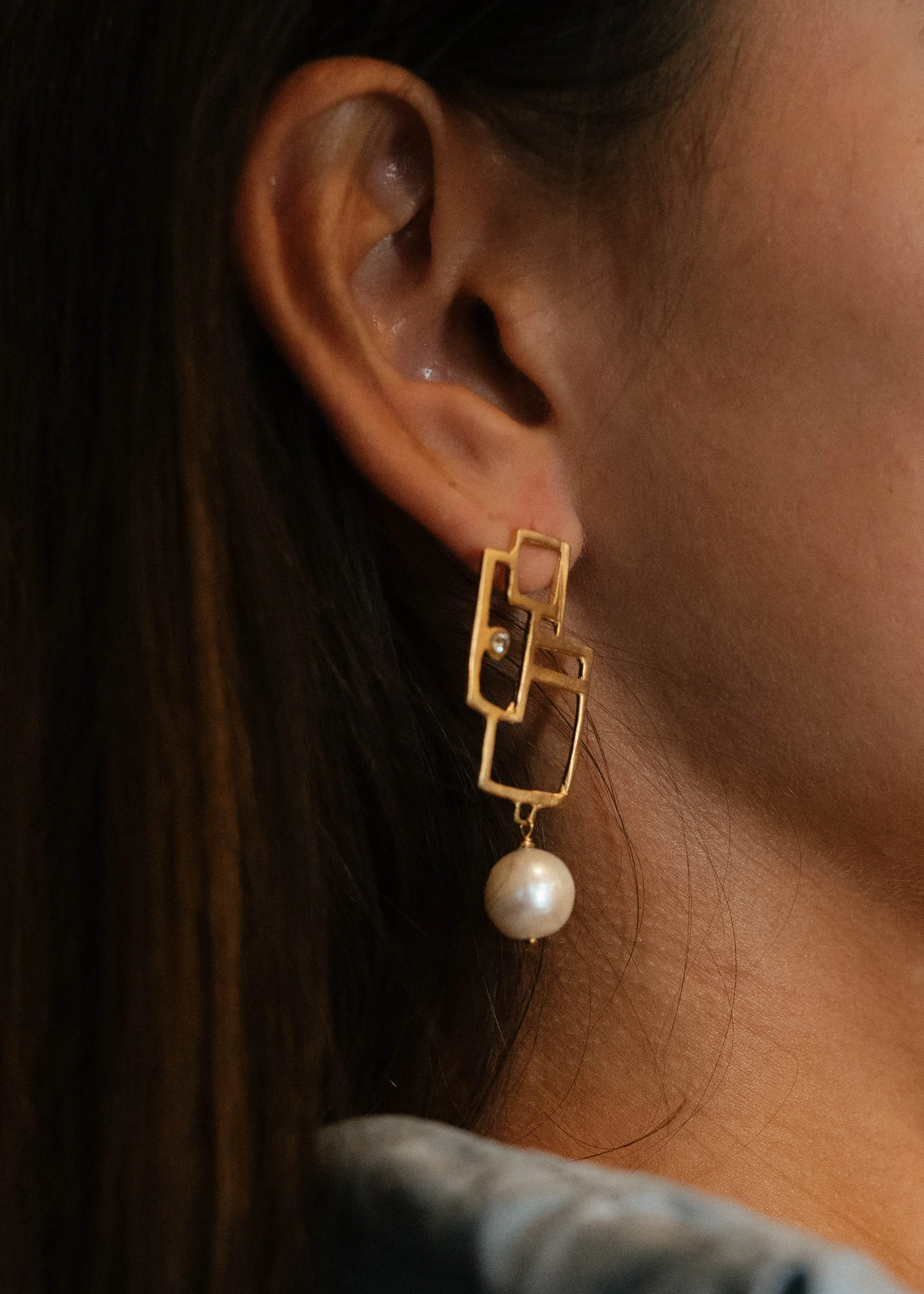 Close-up of a woman's ear wearing a gold geometric earring with a pearl dangling from it.