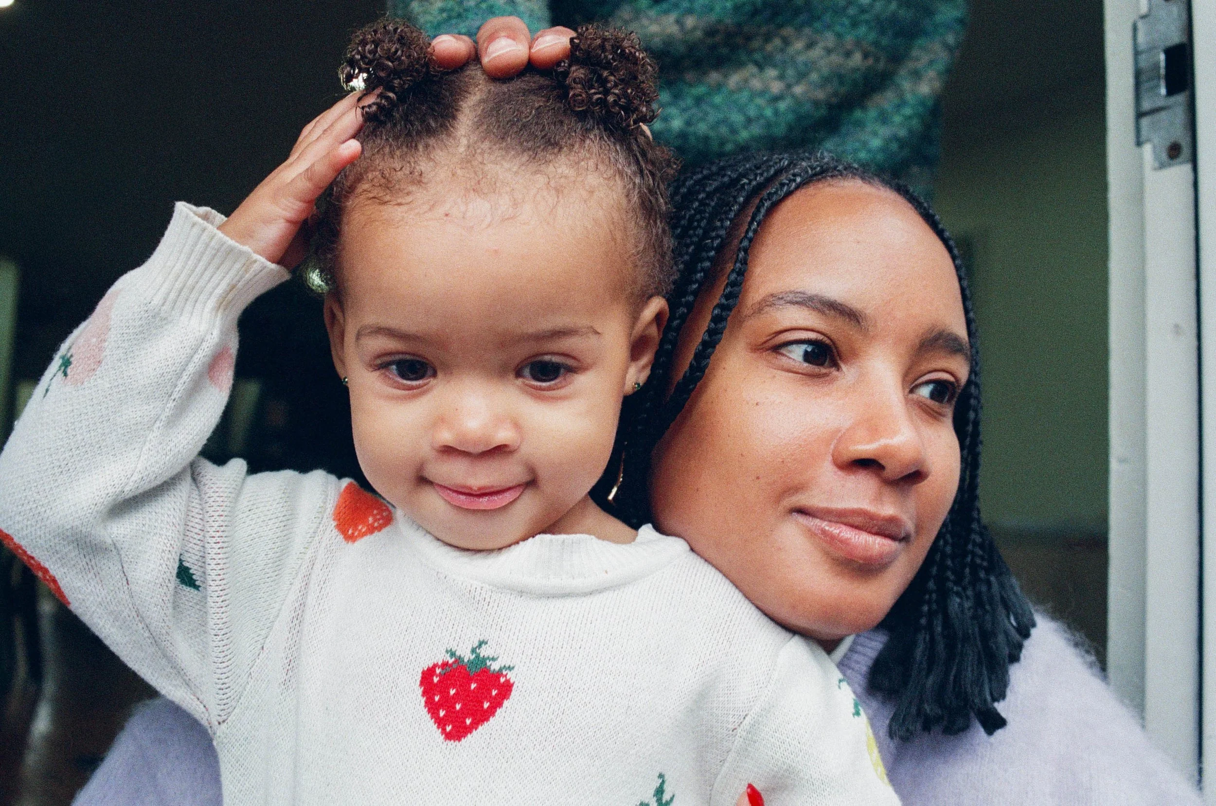 A woman with dark braided hair and a young girl with curly hair and light skin, smiling, close together indoors.