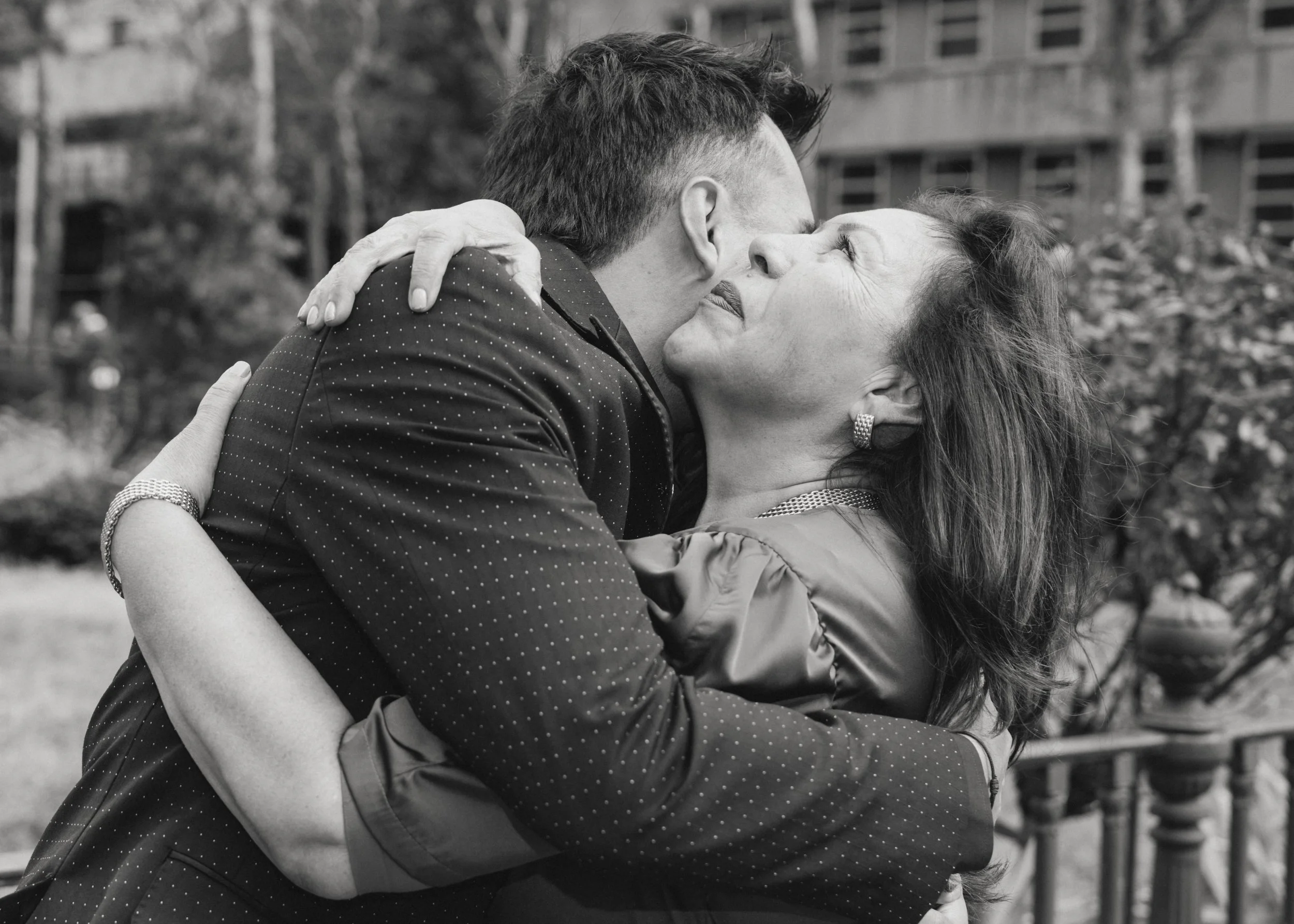 A man and woman embrace and kiss outdoors, with the woman looking up and smiling, in black and white.