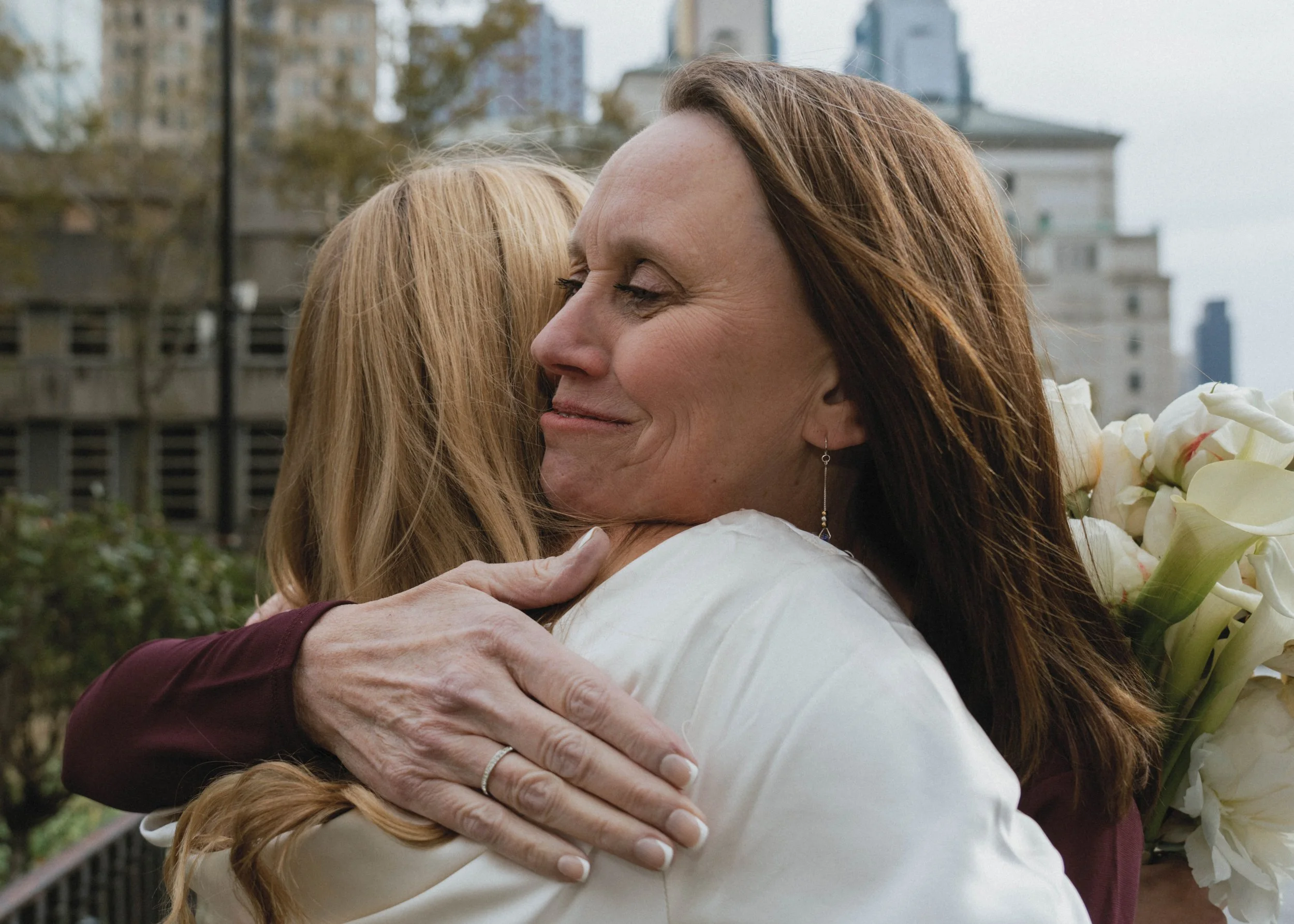 Two women hugging outdoors, one holding a bouquet of white flowers, with buildings and trees in the background.