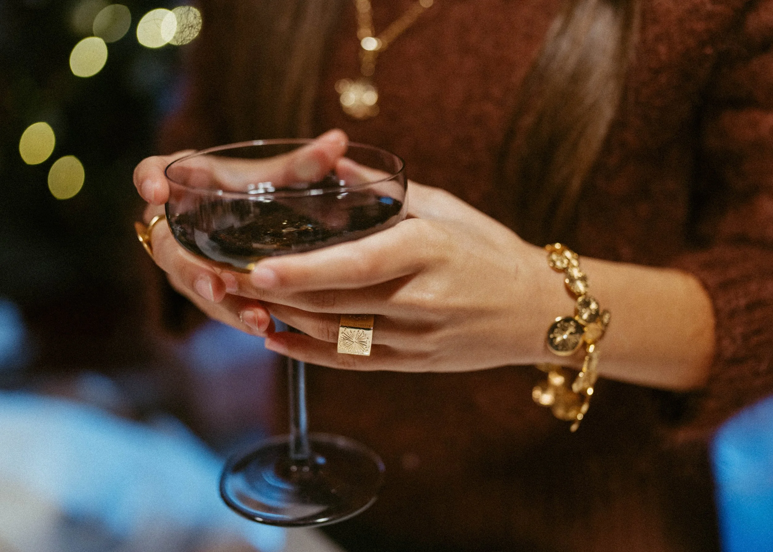 A person holding a glass of red wine with both hands, wearing gold jewelry and a brown sweater, with blurry festive lights in the background.