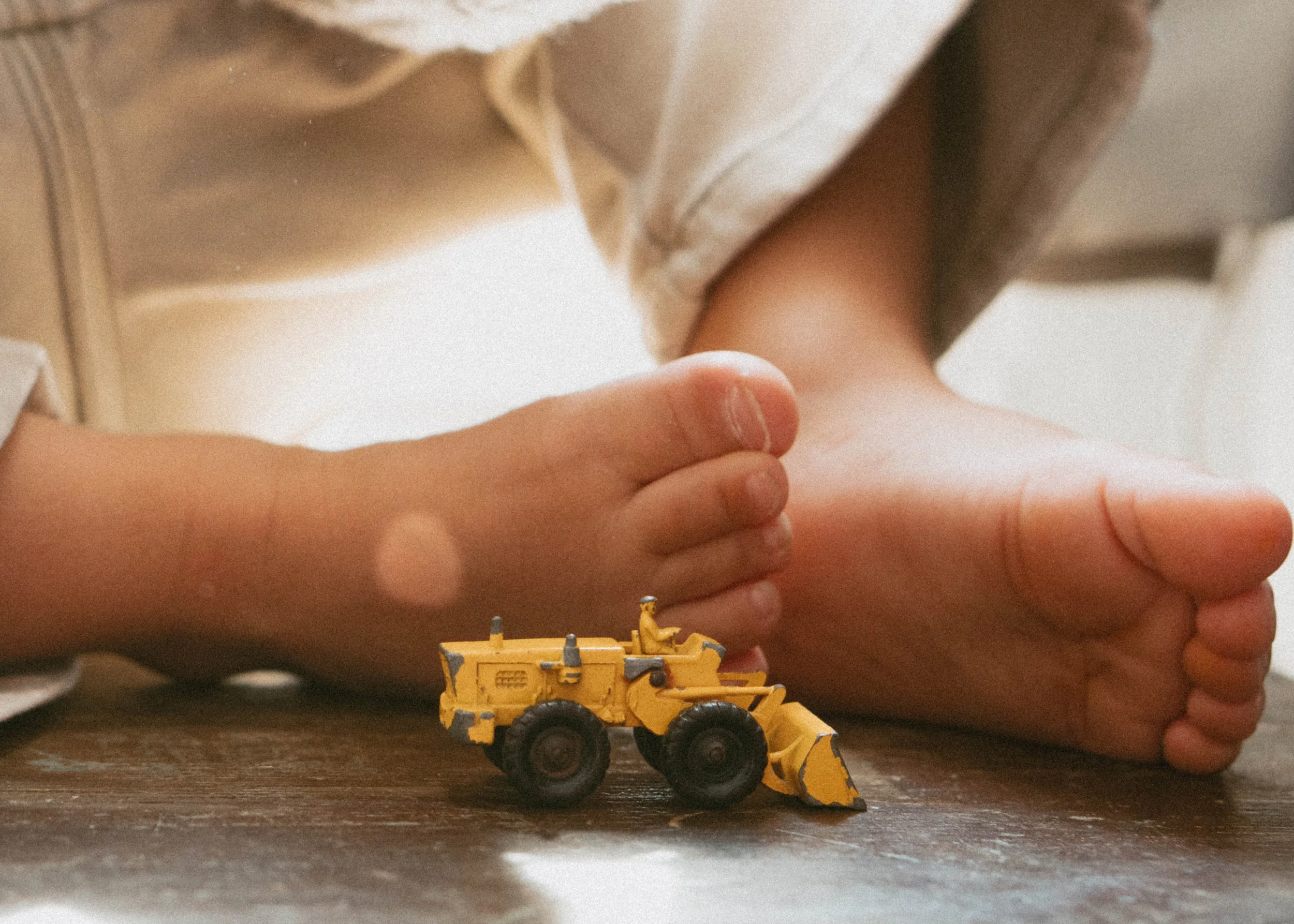 A child's hand is pressed against an adult's hand on a wooden surface, with a small yellow toy bulldozer in the foreground.