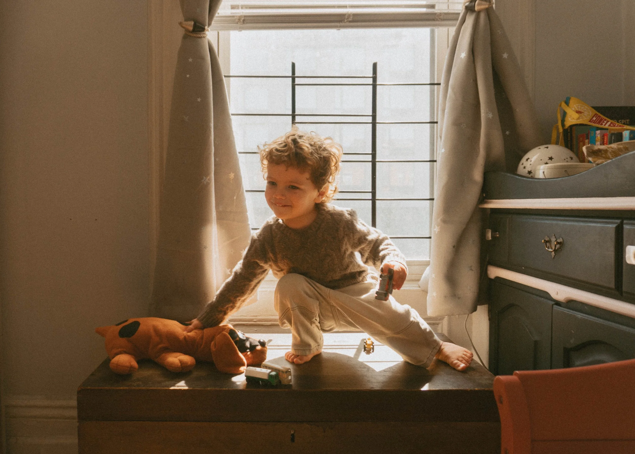 A young child with curly hair playing on a wooden table in a room with a large window. The child is holding a toy car and has a plush dog toy on the table. Sunlight streams through the window, illuminating the scene.