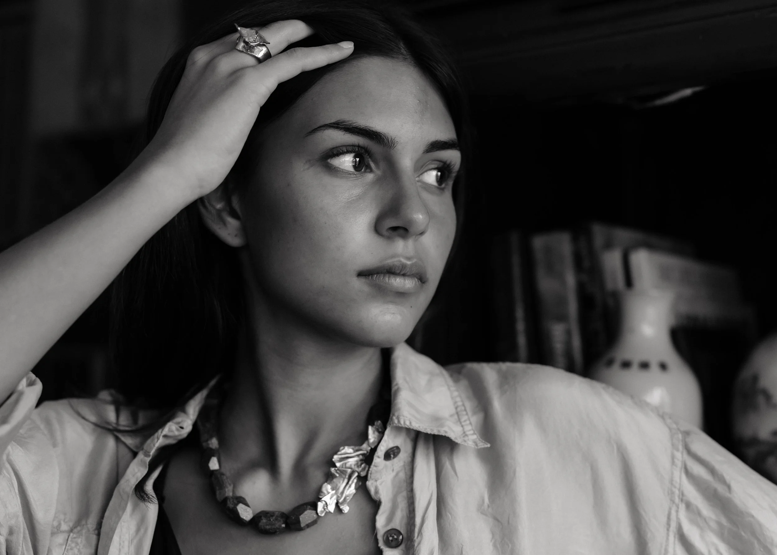 A black-and-white photo of a woman with long dark hair, wearing a light-colored shirt and a chunky necklace, resting her hand on her head while looking off to the side.