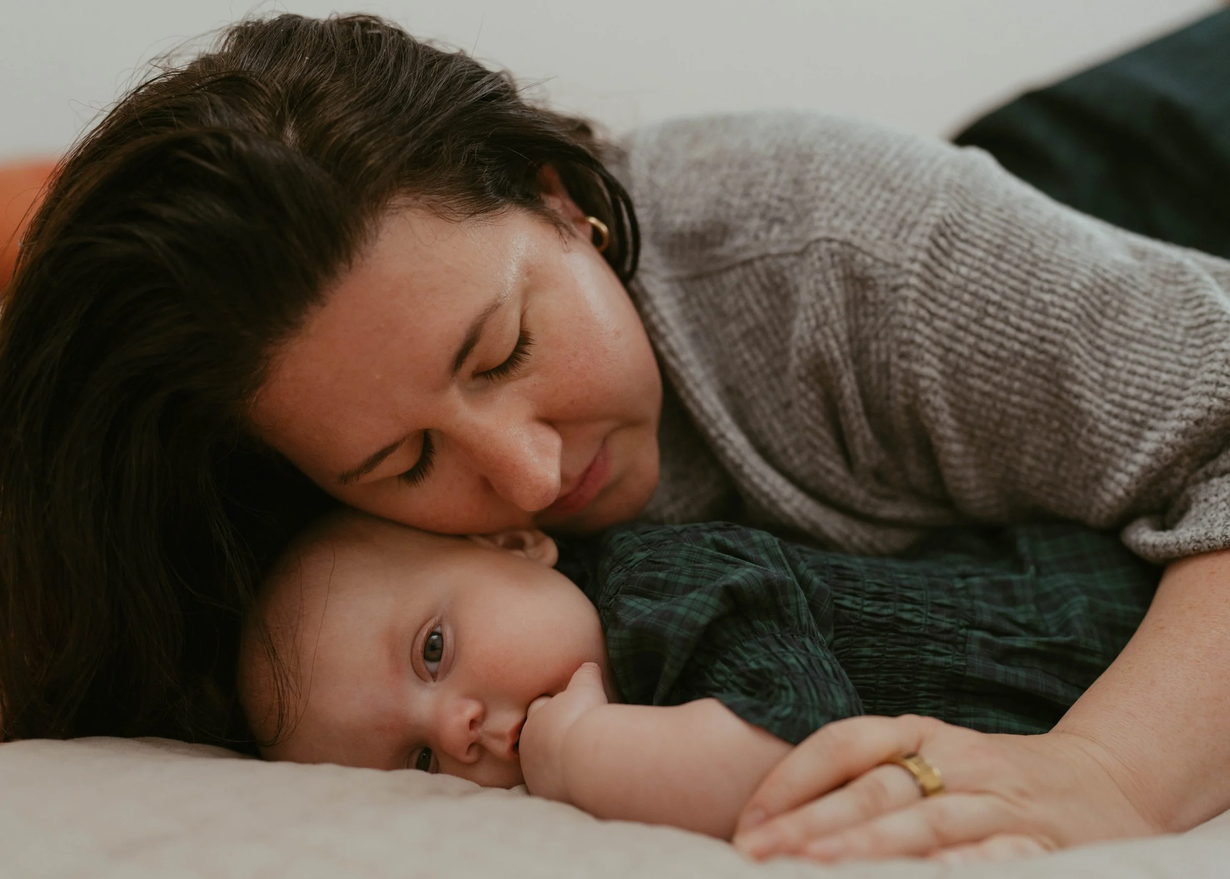 A woman with dark hair and a gray sweater lying on a bed, resting her head close to a baby with light skin and light eyes, who is lying on a bed and sucking their thumb.