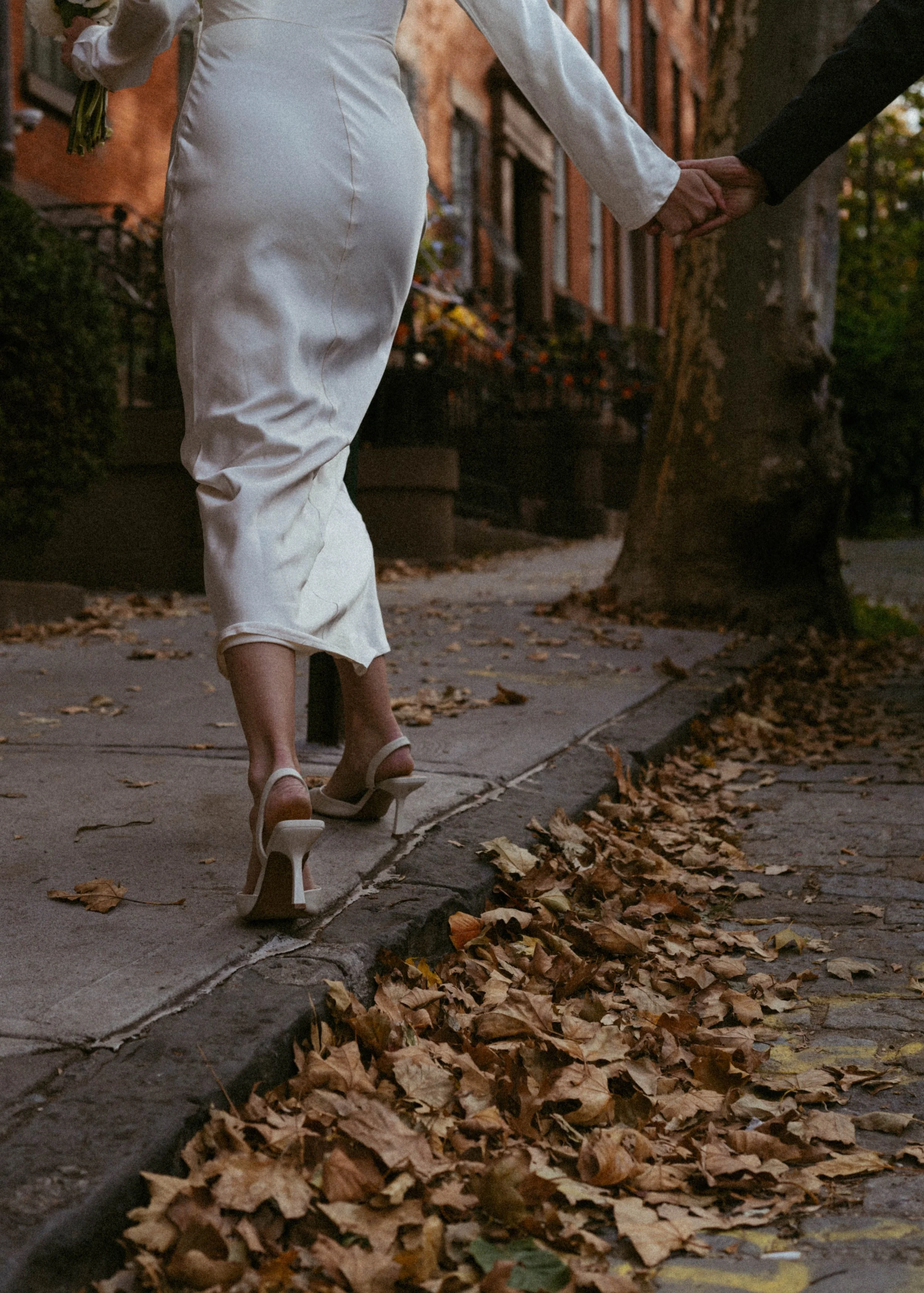 A woman in white high heels and a satin white dress is holding hands with a man in a black suit walking down a sidewalk lined with fallen autumn leaves, trees, and residential buildings.