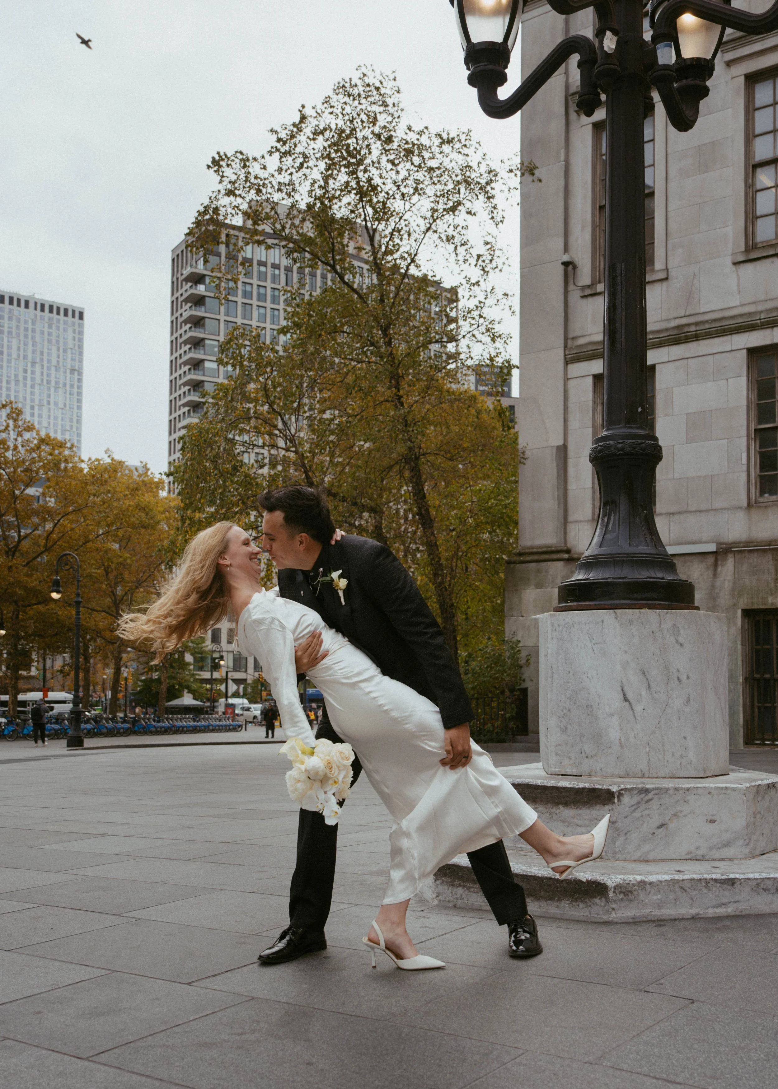 A newlywed couple is dancing in an urban park, with the groom dipping the bride and both smiling. The city skyline and autumn trees are in the background.