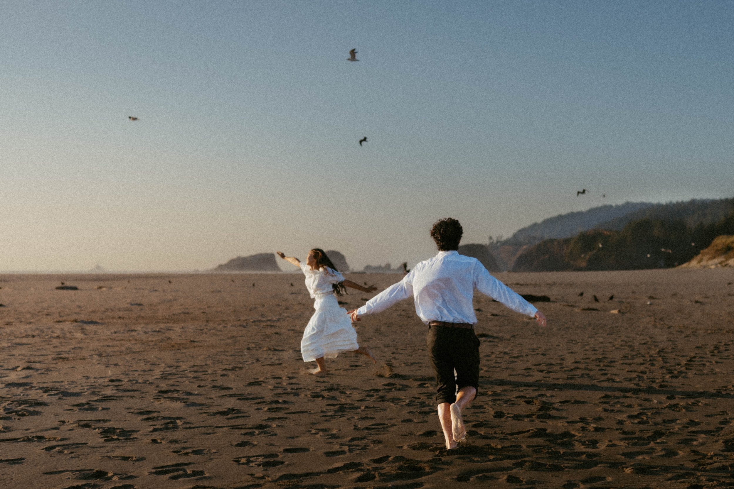 A couple running on a beach at sunset with a tan sandy shoreline, distant hills, and a blue sky with a few seagulls flying overhead.