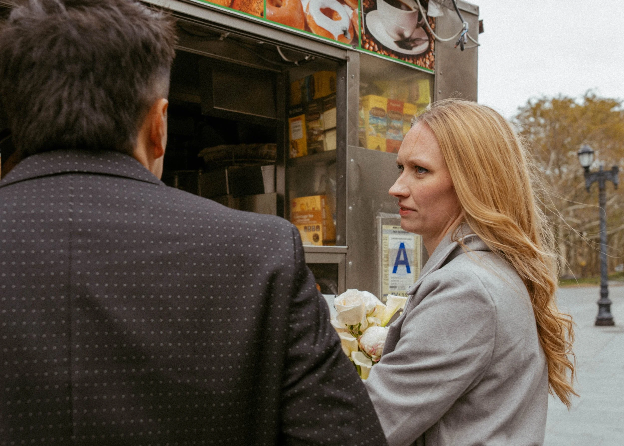 A woman with long, wavy red hair holding white roses, facing a man in a dark jacket with small white dots, near a food cart outdoors.
