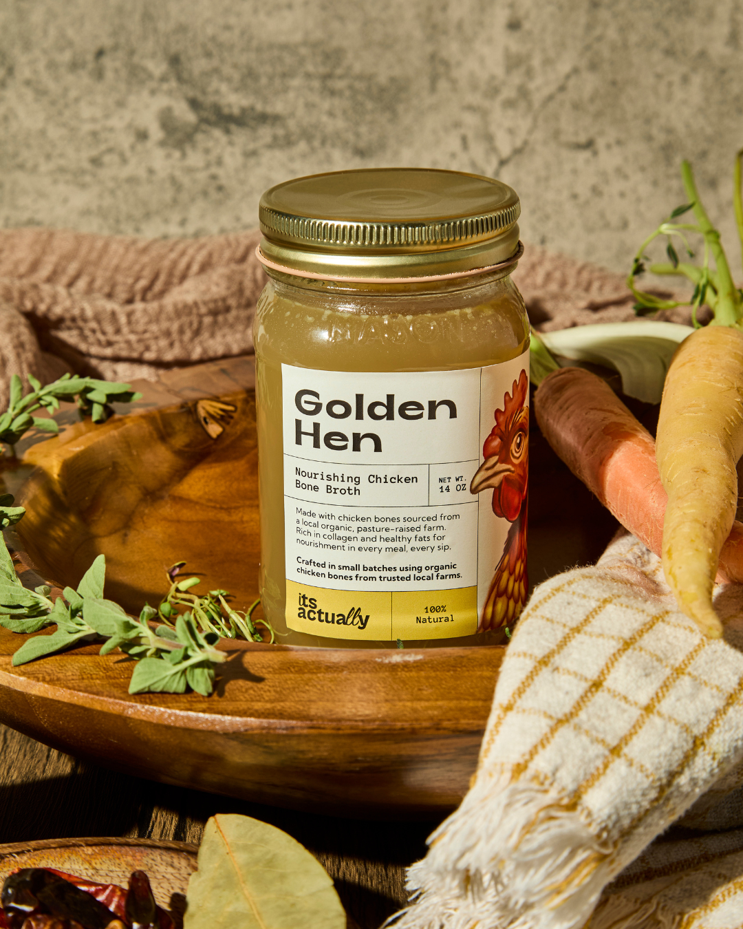 A jar of Golden Hen nourishing chicken bone broth on a wooden table surrounded by fresh carrots, herbs, and a cloth towel.